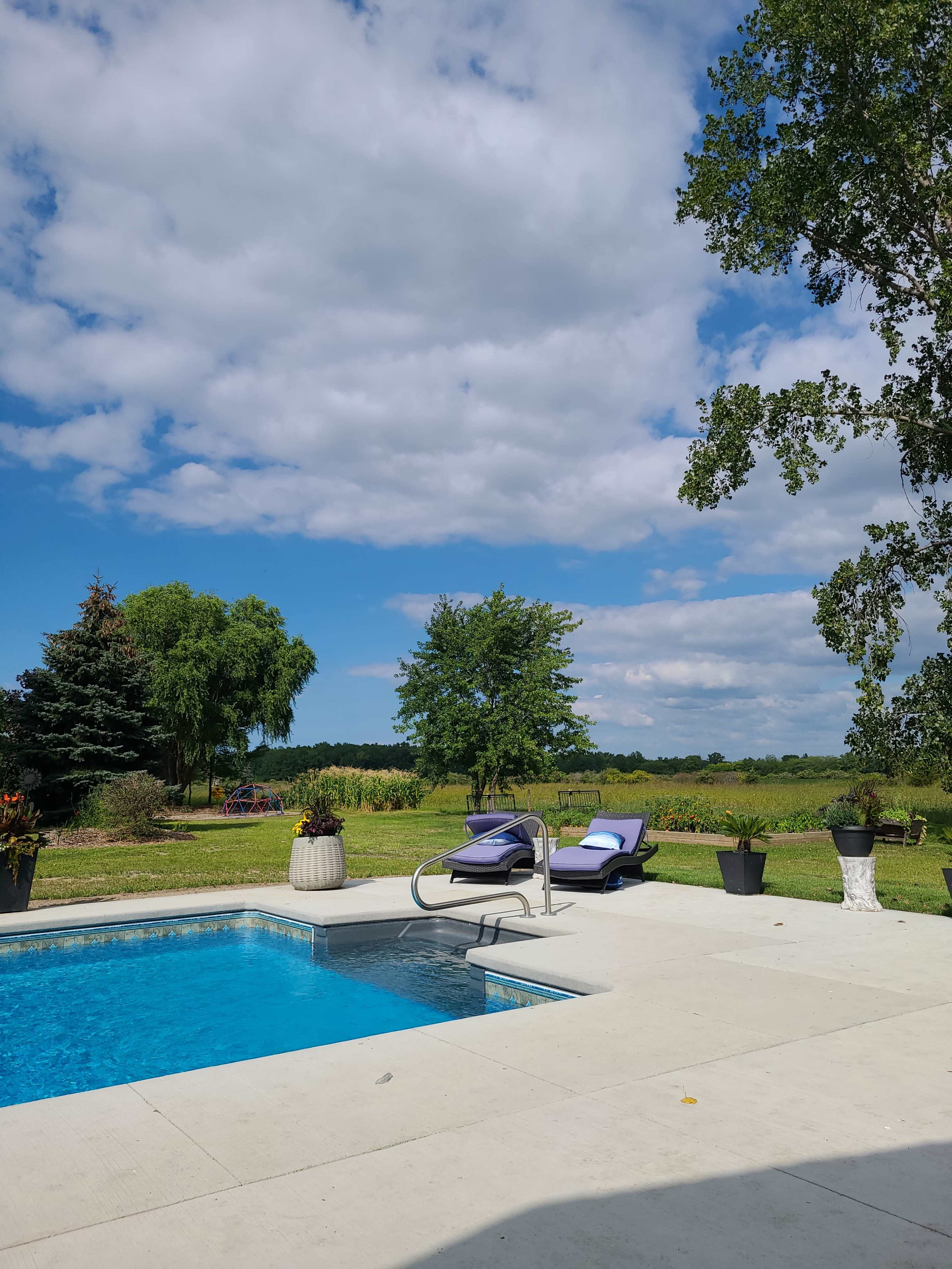 A swimming pool with two lounge chairs is surrounded by a grass field and trees under a partly cloudy sky.