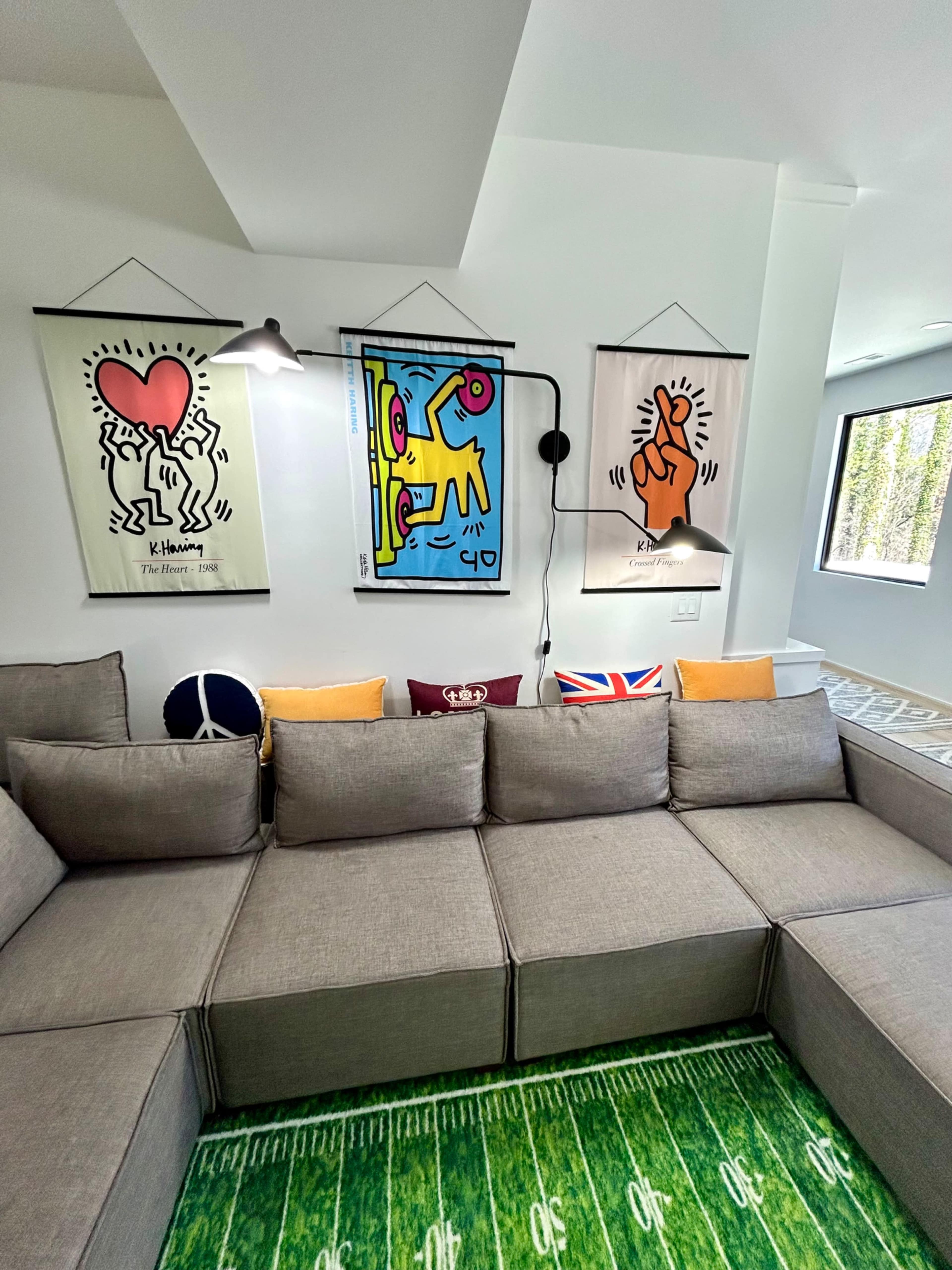 The image shows a modern living space featuring a gray sectional sofa adorned with decorative pillows and three vibrant art pieces hanging on the wall.