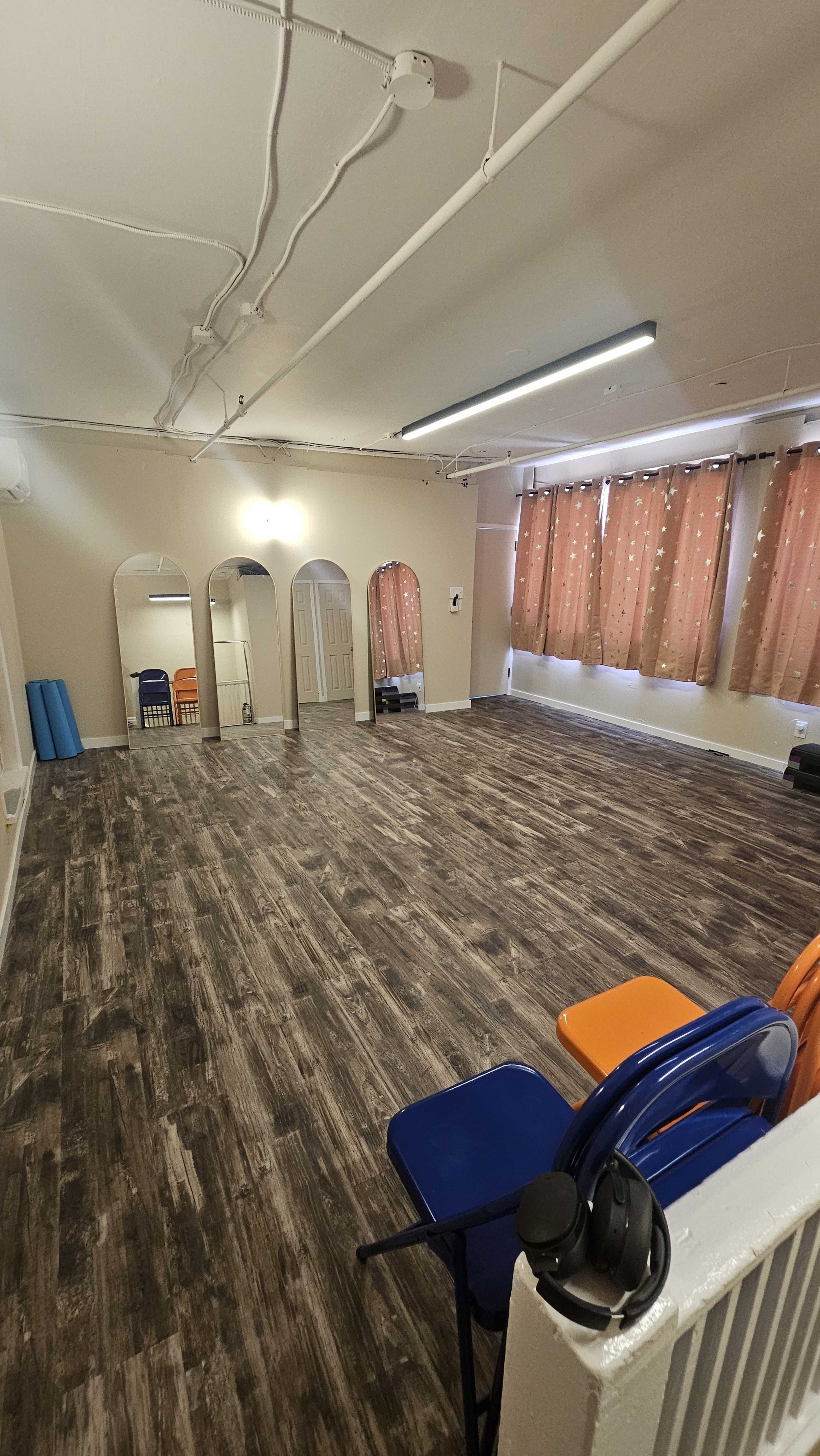The image shows a spacious room with wooden flooring, featuring several arches, orange and blue chairs, and large windows covered with polka-dotted curtains.