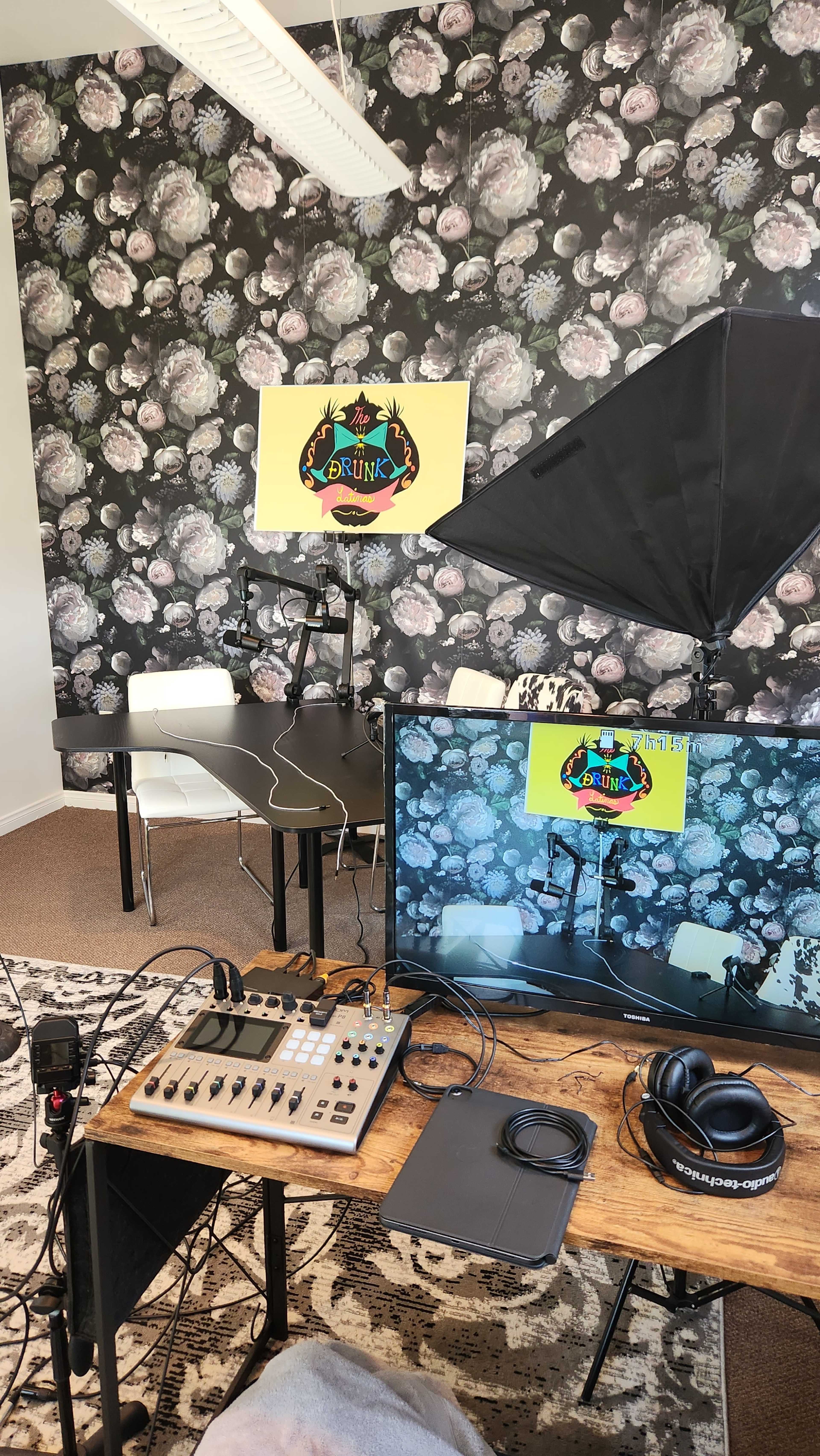 A media studio setup featuring a floral wallpaper backdrop, a large display screen, a microphone, a mixer, headphones, and lighting equipment arranged around a table.
