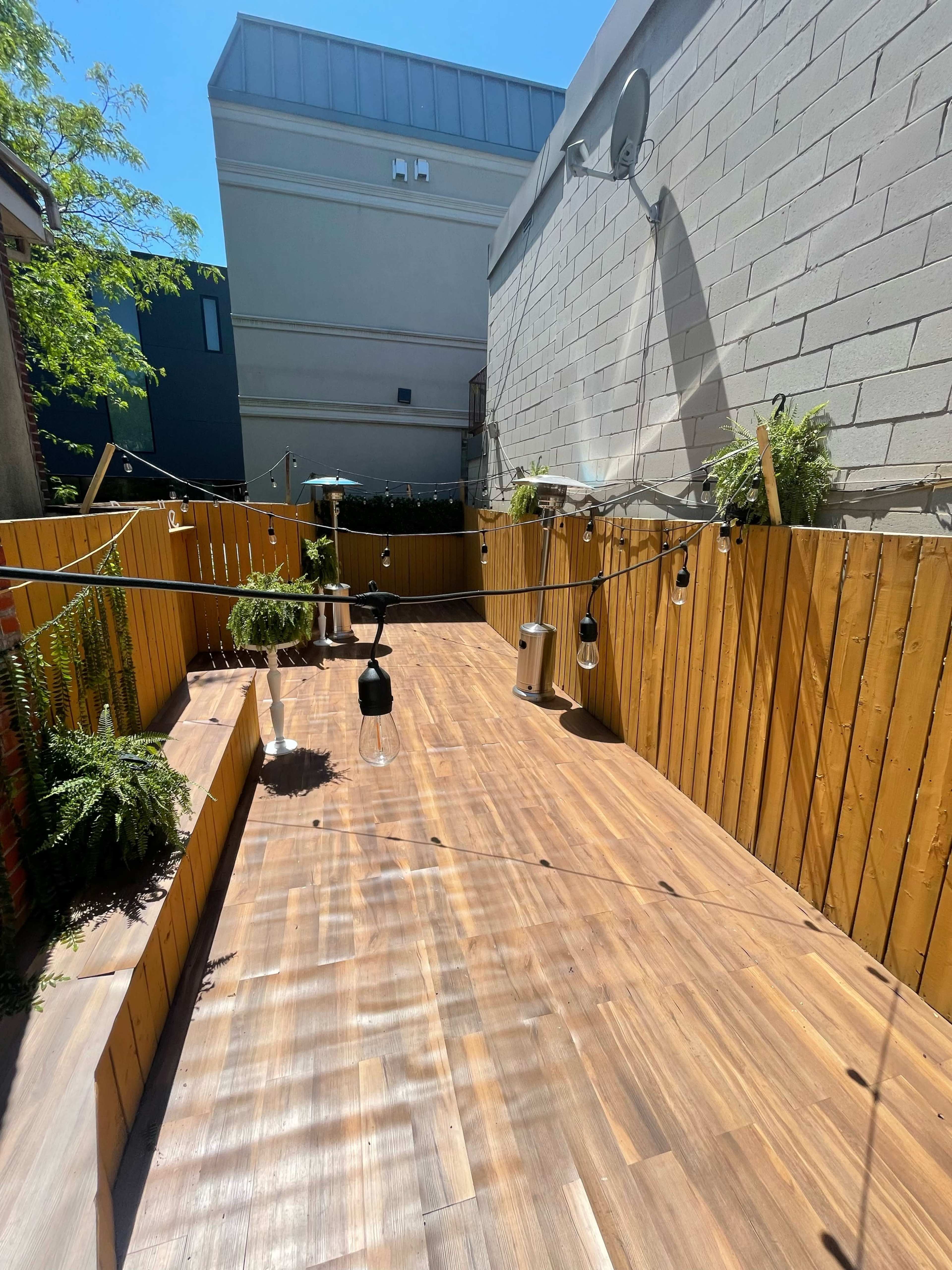 A wooden outdoor space with hanging lights and plants against a backdrop of a brick and gray wall, under a clear blue sky.