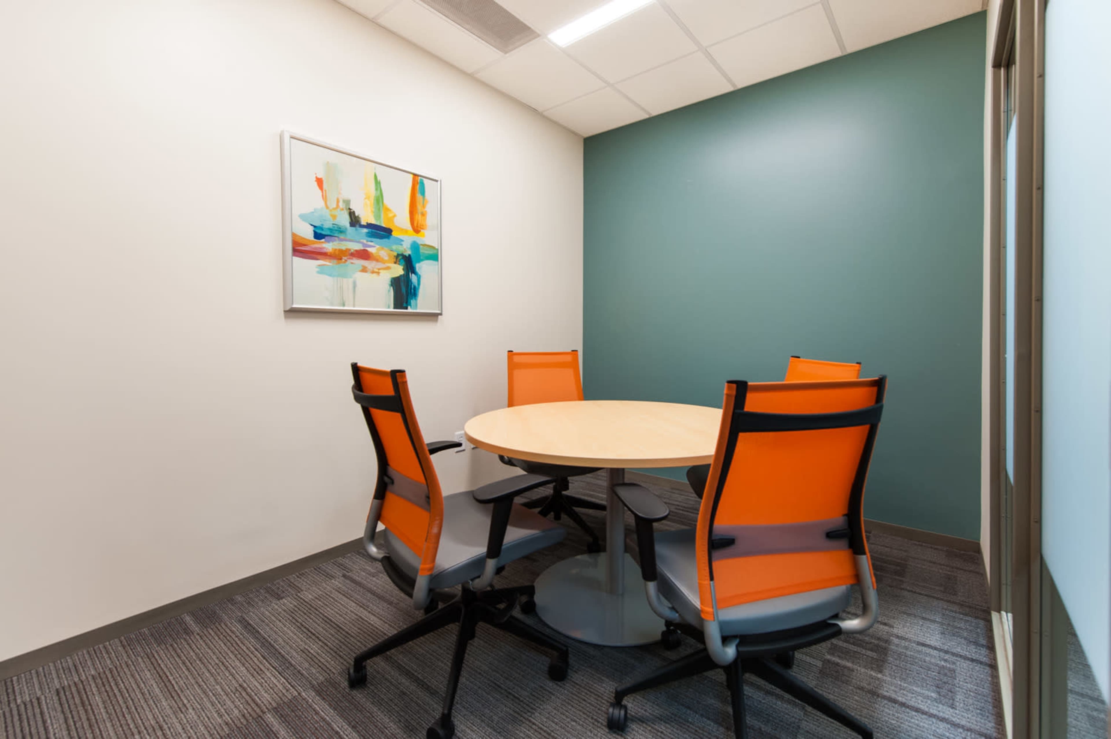 A small meeting room features a round wooden table surrounded by four orange and gray chairs, with a colorful artwork hanging on the wall.