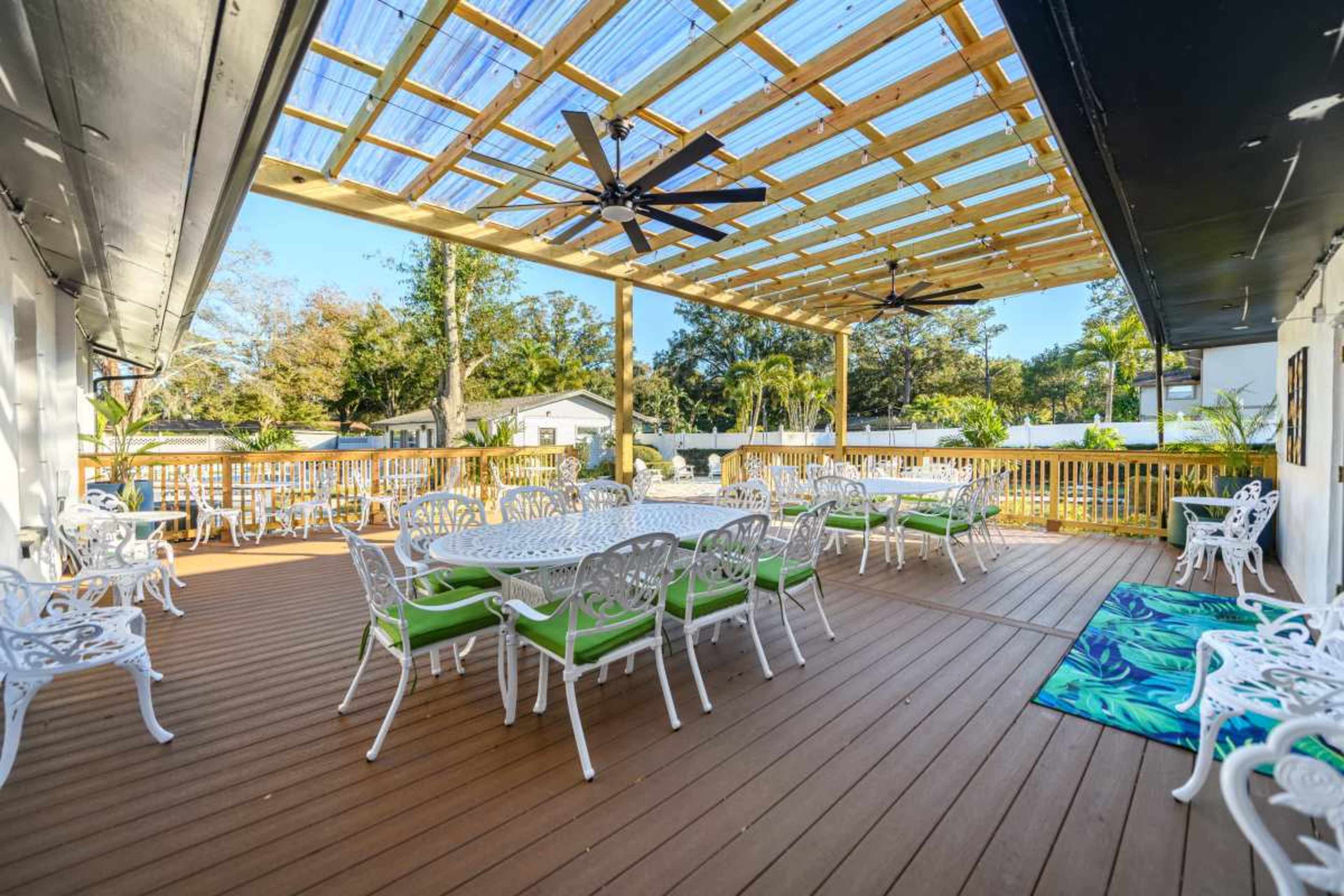 The image shows a spacious outdoor deck with multiple white tables and chairs, a wooden ceiling structure, and ceiling fans, surrounded by greenery.