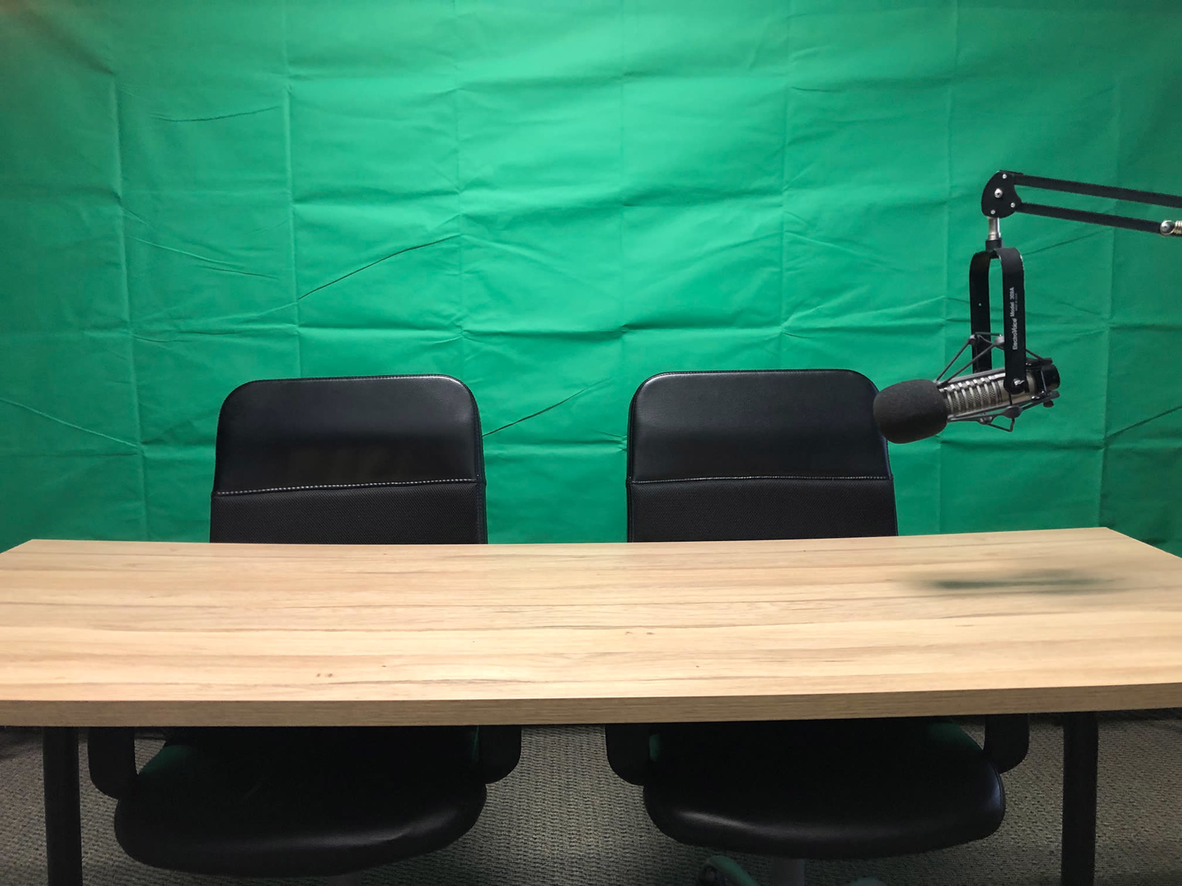 The image shows a solid wood table with two black chairs in front of a bright green backdrop, along with a microphone on an adjustable arm.