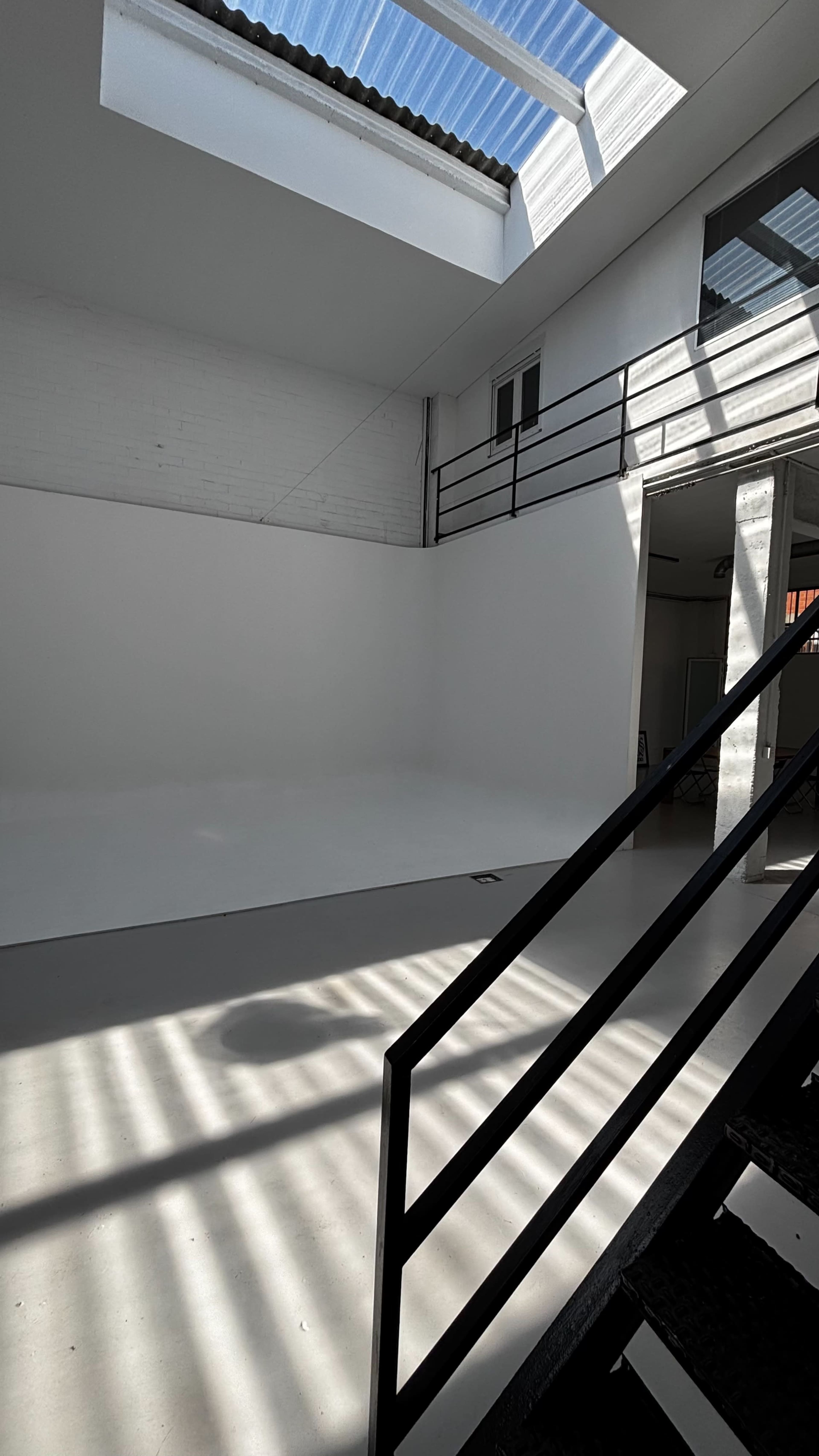 The image shows a bright, minimalist interior space with a white wall, a skylight, and a staircase in the foreground.
