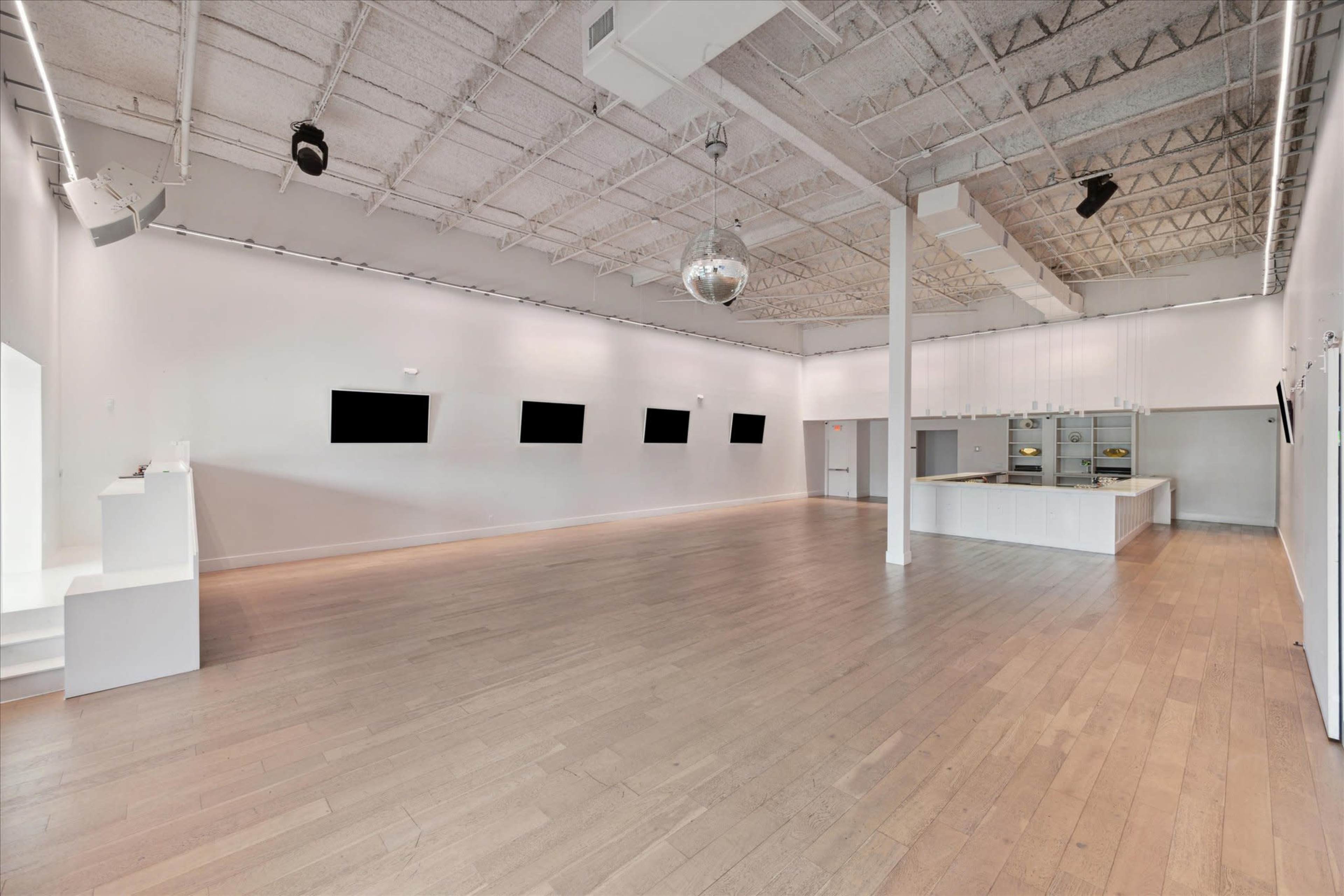 A spacious, white-walled gallery with a wooden floor, featuring a modern kitchen area and several wall-mounted screens.