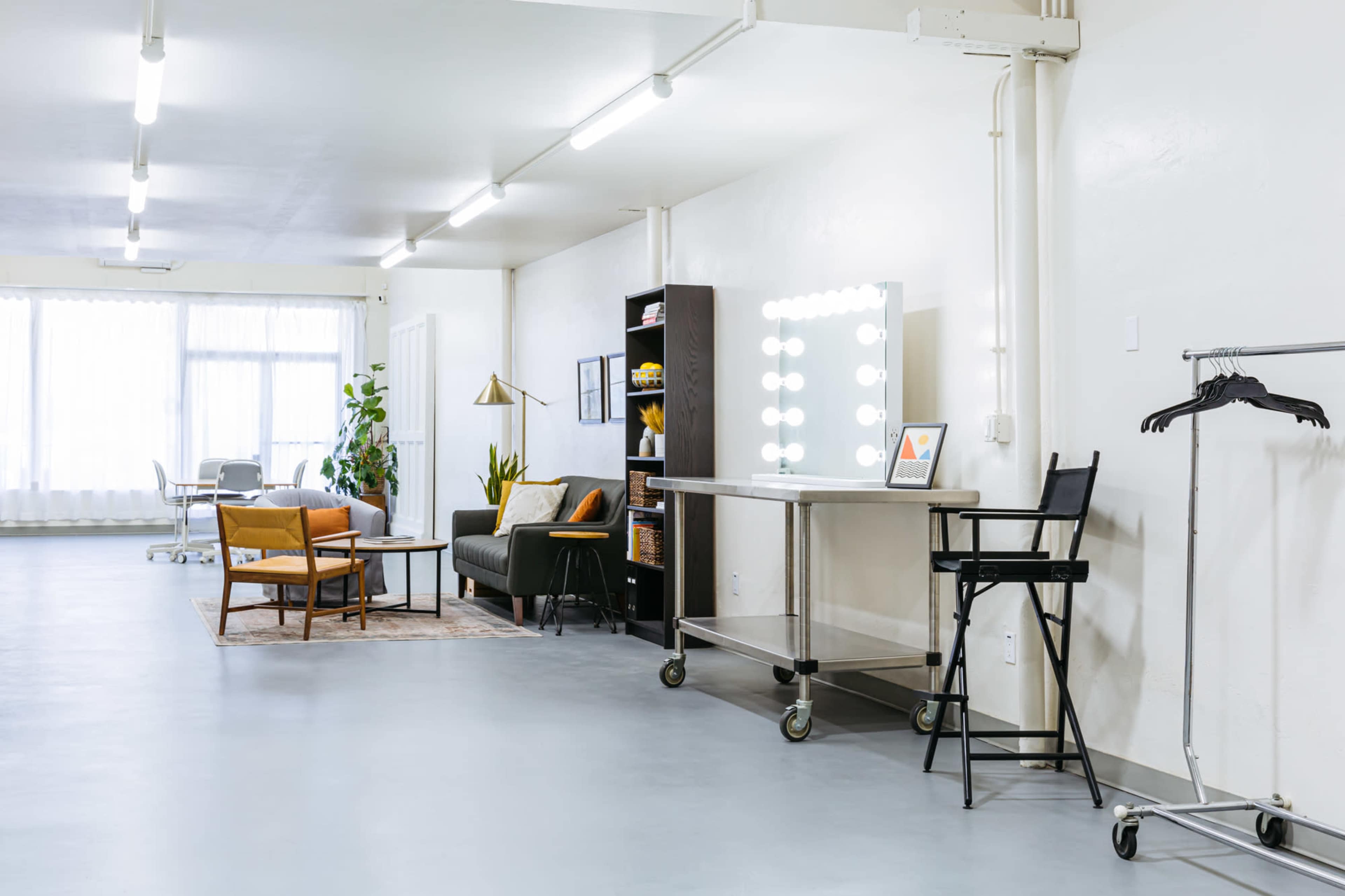 A spacious, minimalist room with white walls, bright lighting, a seating area with a sofa and chairs, a large mirror with lights, and a clothing rack.