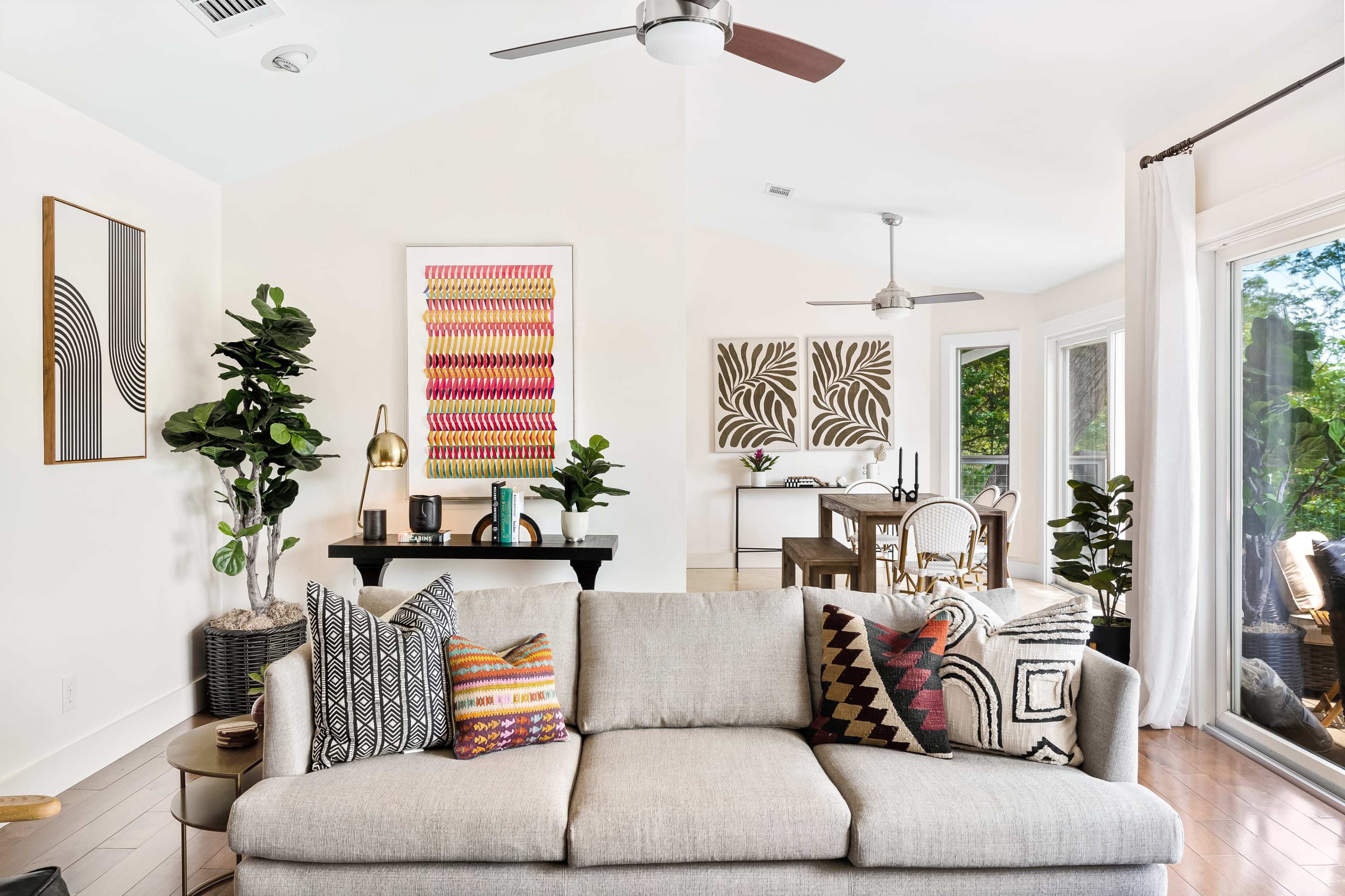 A modern living room features a light gray sofa adorned with patterned pillows, framed art on the walls, and a dining area in the background with a table and chairs.