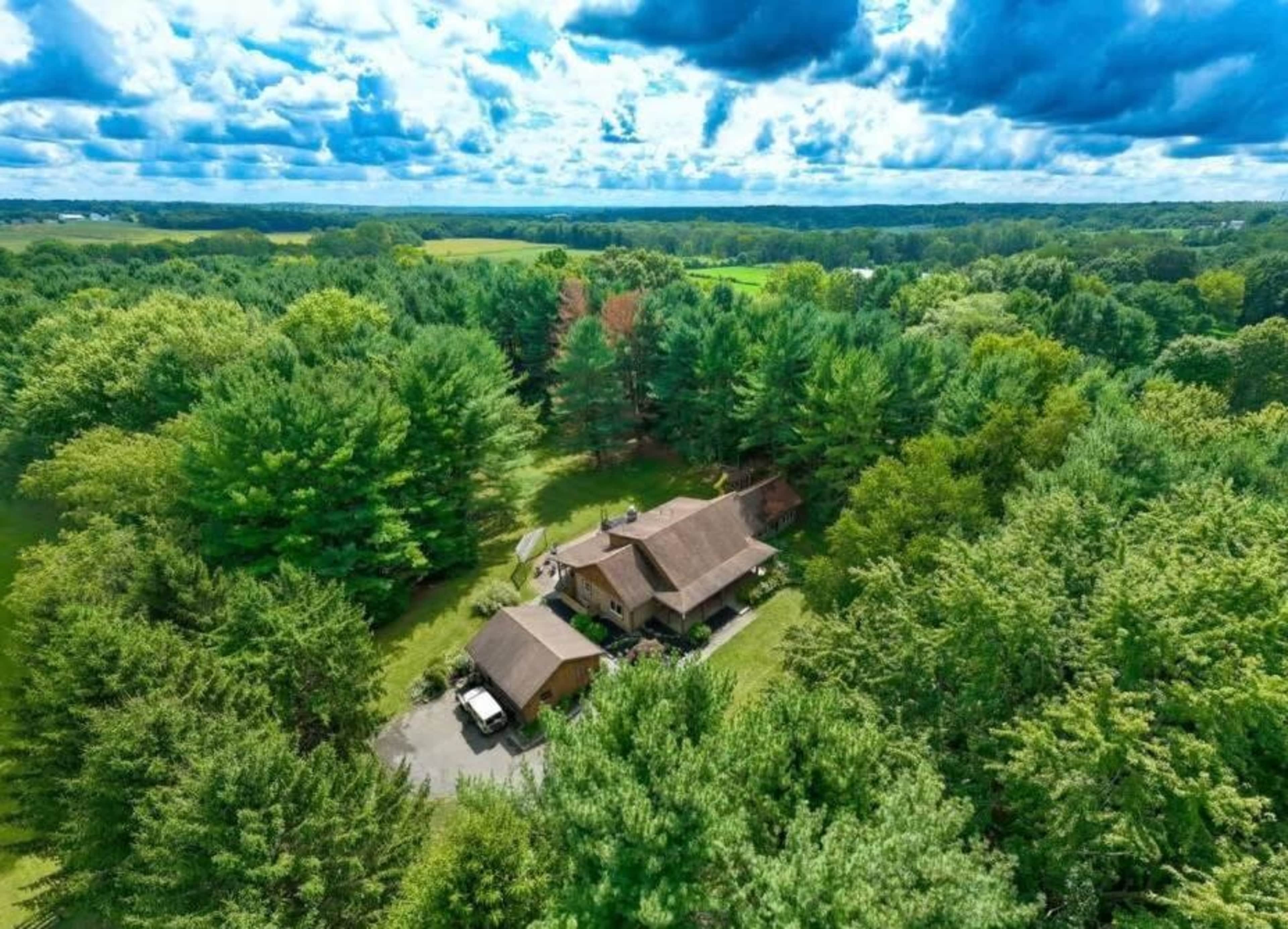 An aerial view shows a house surrounded by dense trees and greenery under a partly cloudy sky.