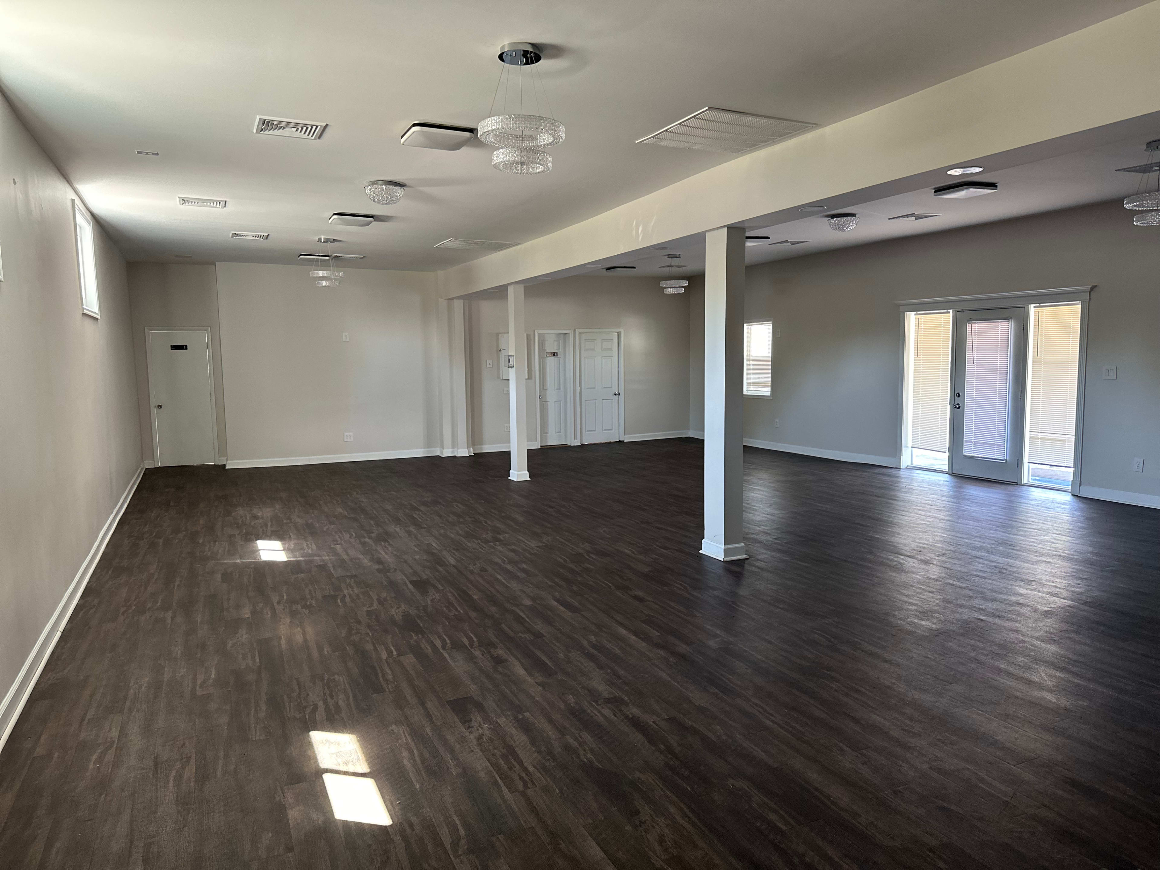 An empty, well-lit interior space with a dark wooden floor and large windows, featuring white walls and several light fixtures.