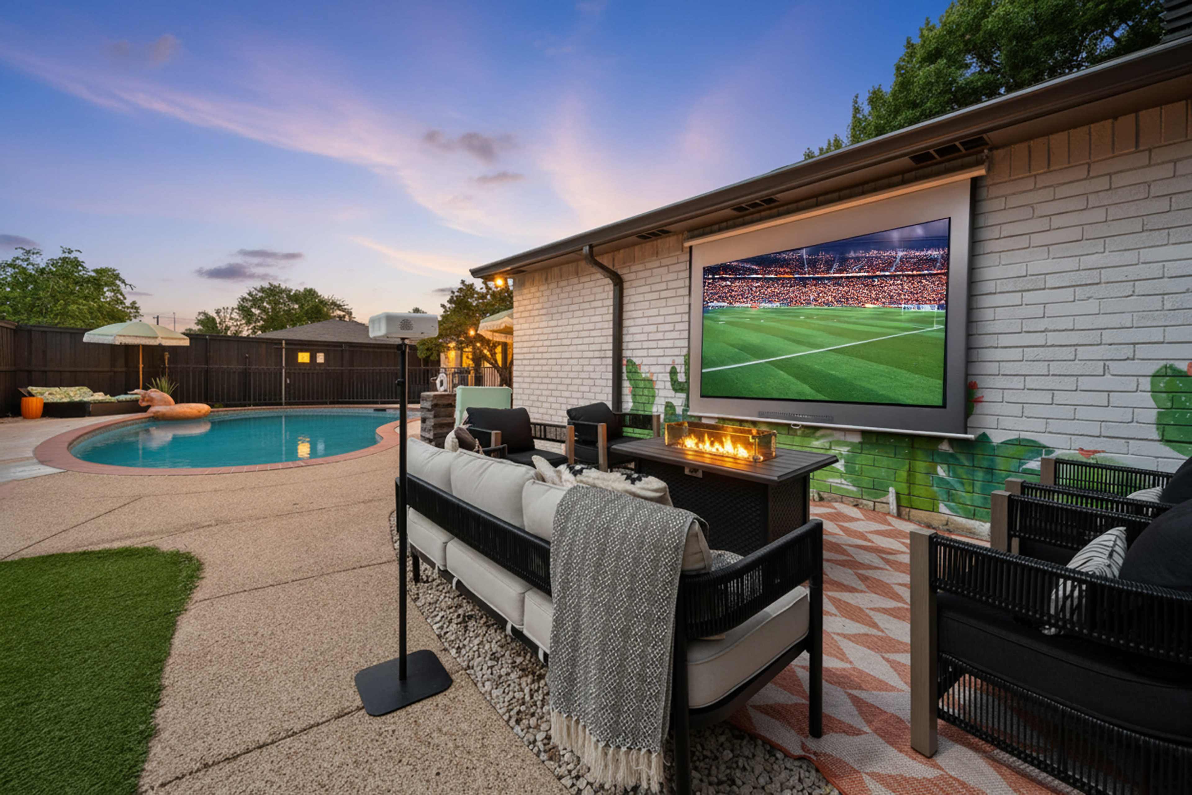A stylish outdoor seating area with a fire pit and a projector screen displaying a soccer match, adjacent to a swimming pool.