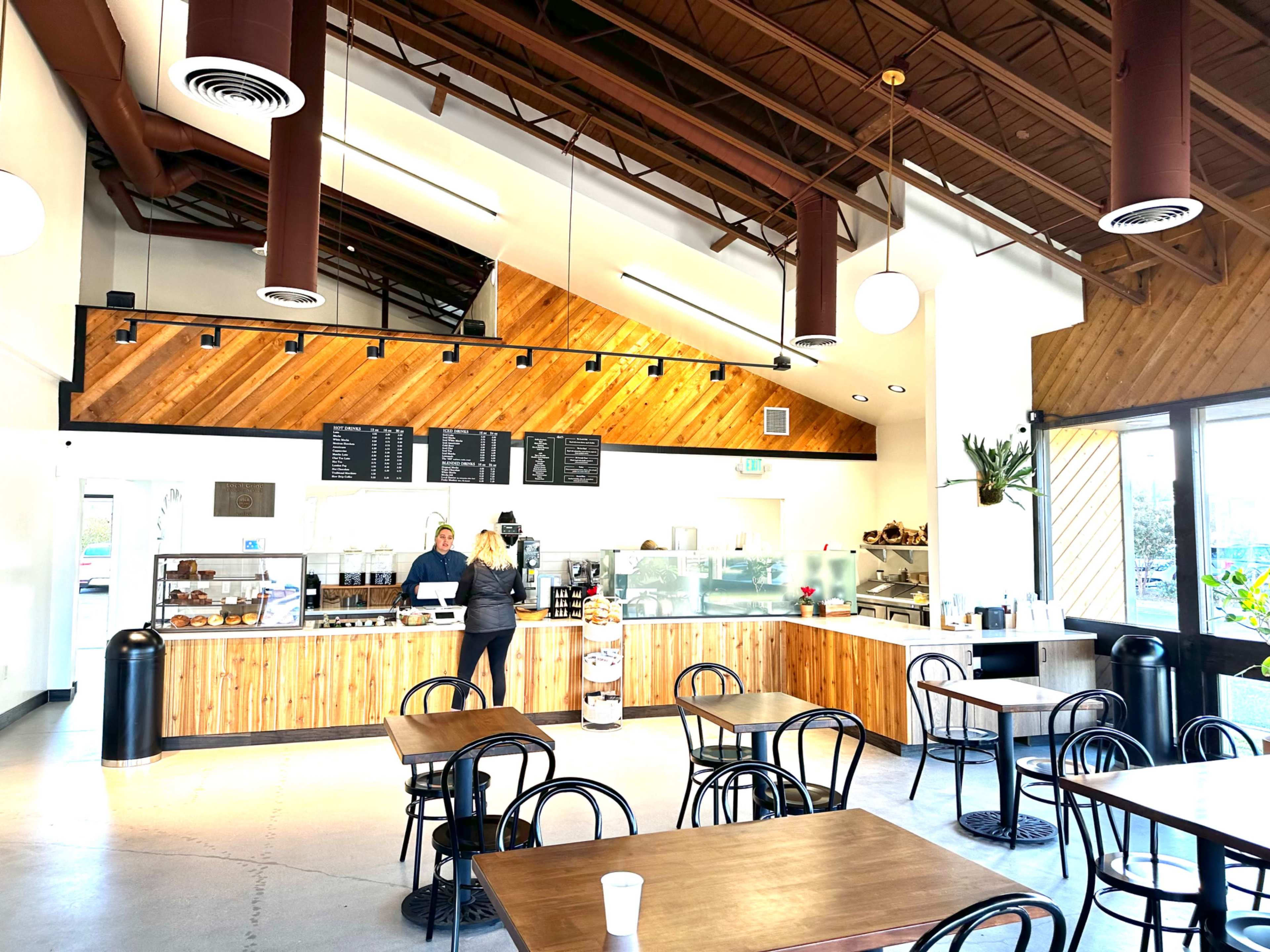 A cafe features wooden counter and tables, with a barista attending to customers in a bright, open space with high ceilings and large windows.