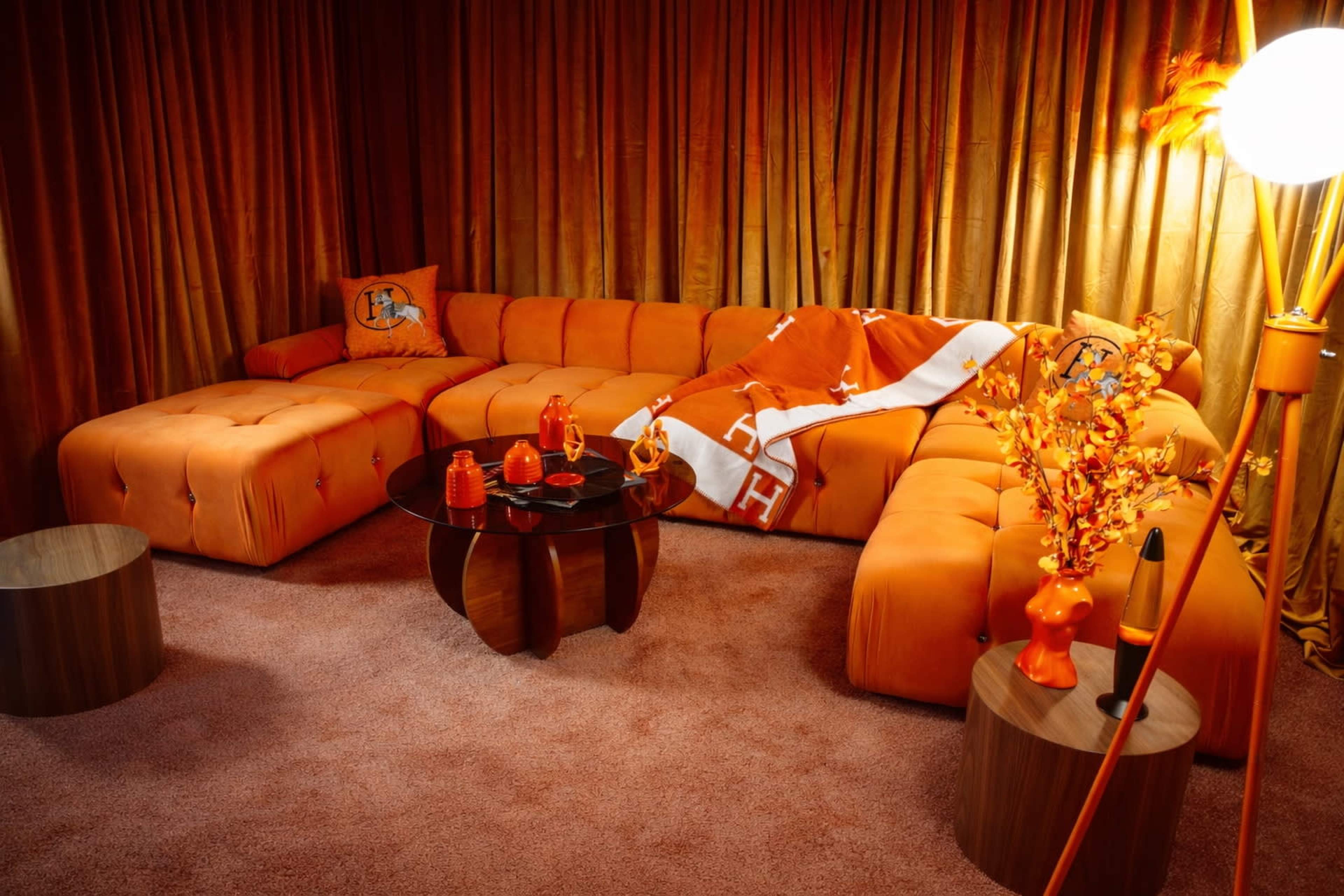 The scene features an orange-themed living room with a plush sectional sofa, a round coffee table, decorative orange objects, and curtains that match the overall color scheme.
