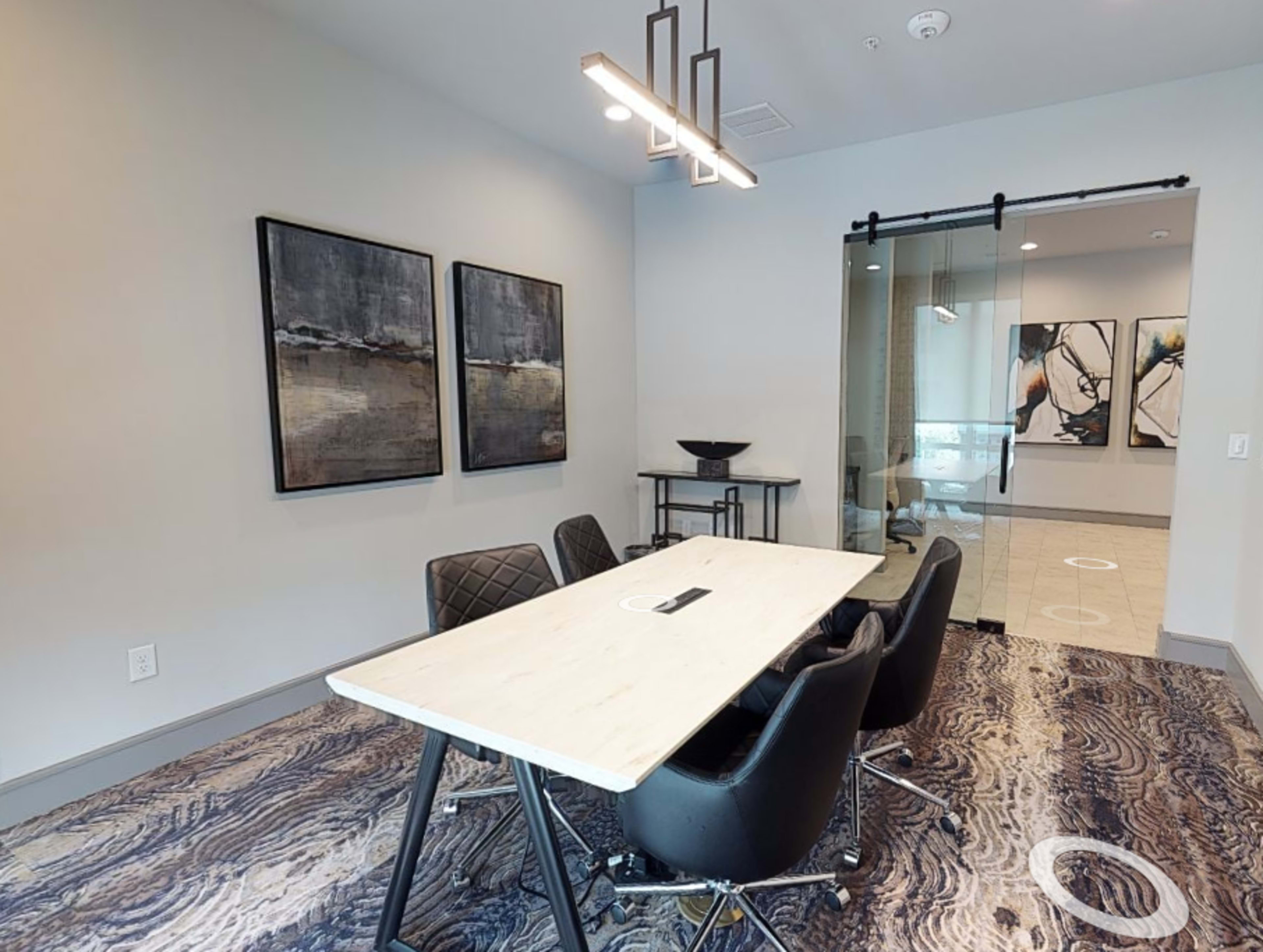 The image shows a modern conference room featuring a marble-topped table with four black chairs, two abstract paintings on the wall, and a glass door leading to another area.
