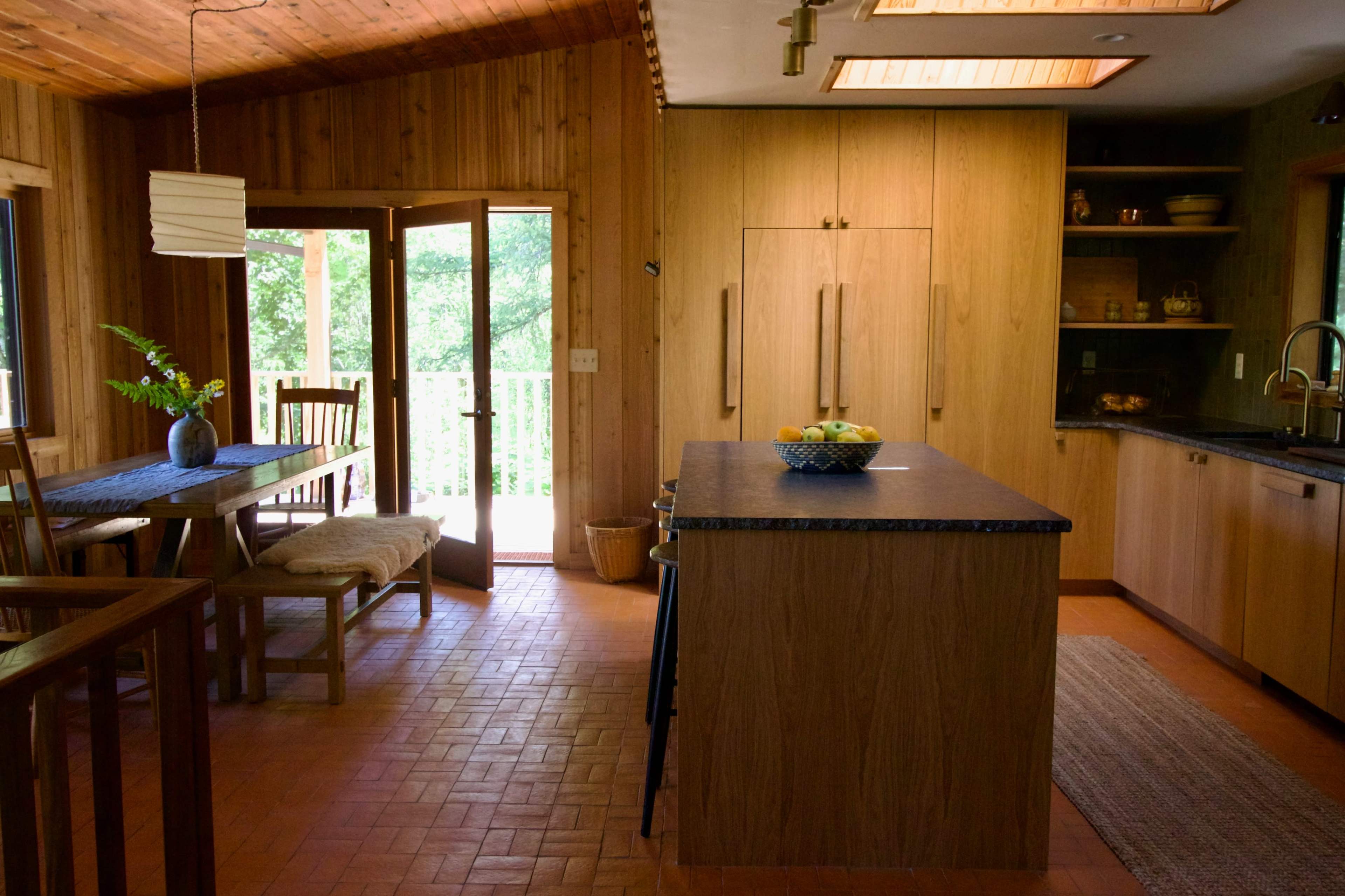 The image shows a warm wood-finished kitchen and dining area with a large island, a wooden dining table, and open doors leading to a deck.