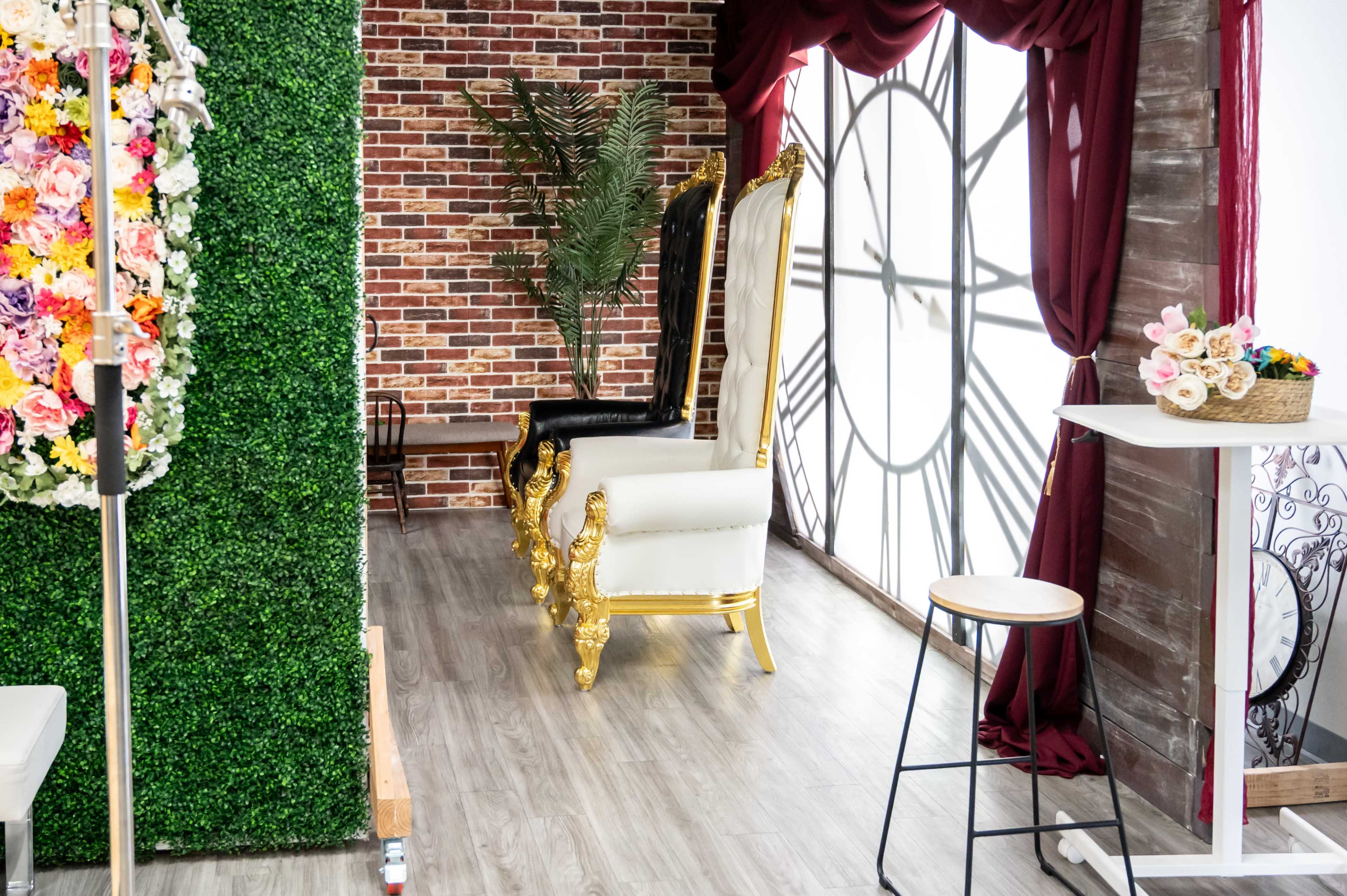 The image shows a decorated indoor space featuring a large black and gold throne, a backdrop of brick wall, greenery, and elegant curtains.