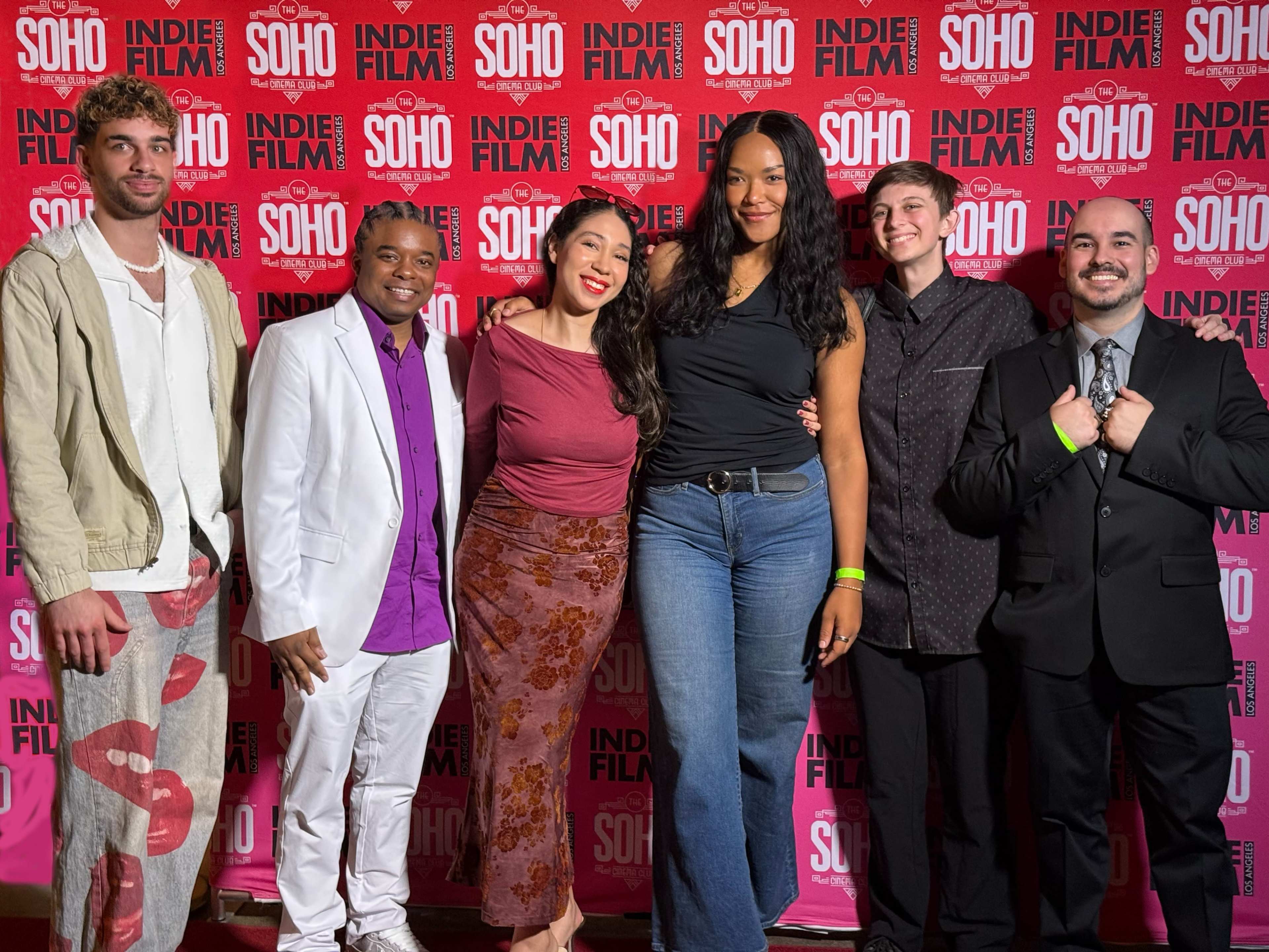 A group of six individuals stands together on a red carpet at an event, posing in front of a branded backdrop.