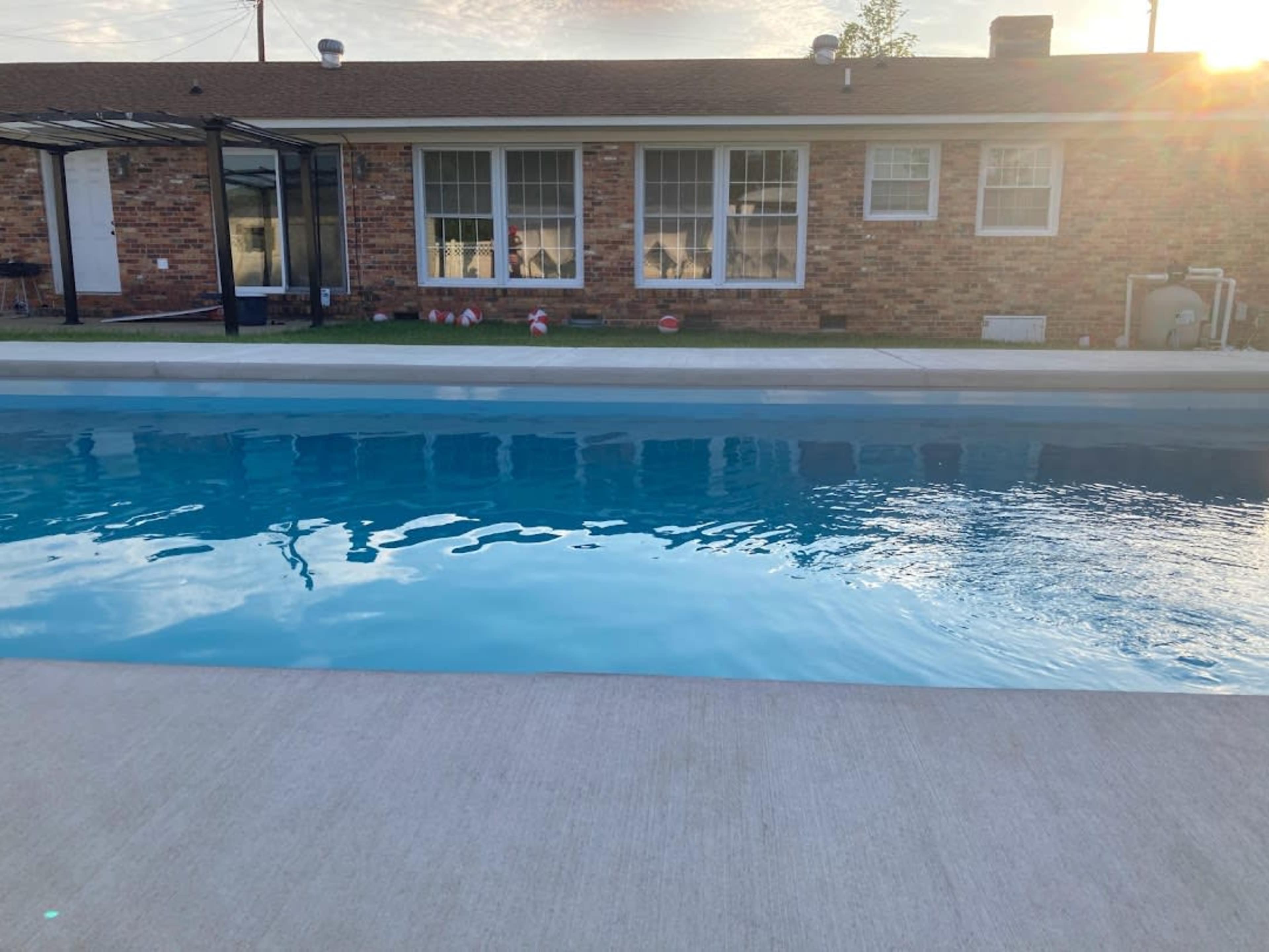 A clear swimming pool with a concrete deck in front of a brick house featuring large windows.