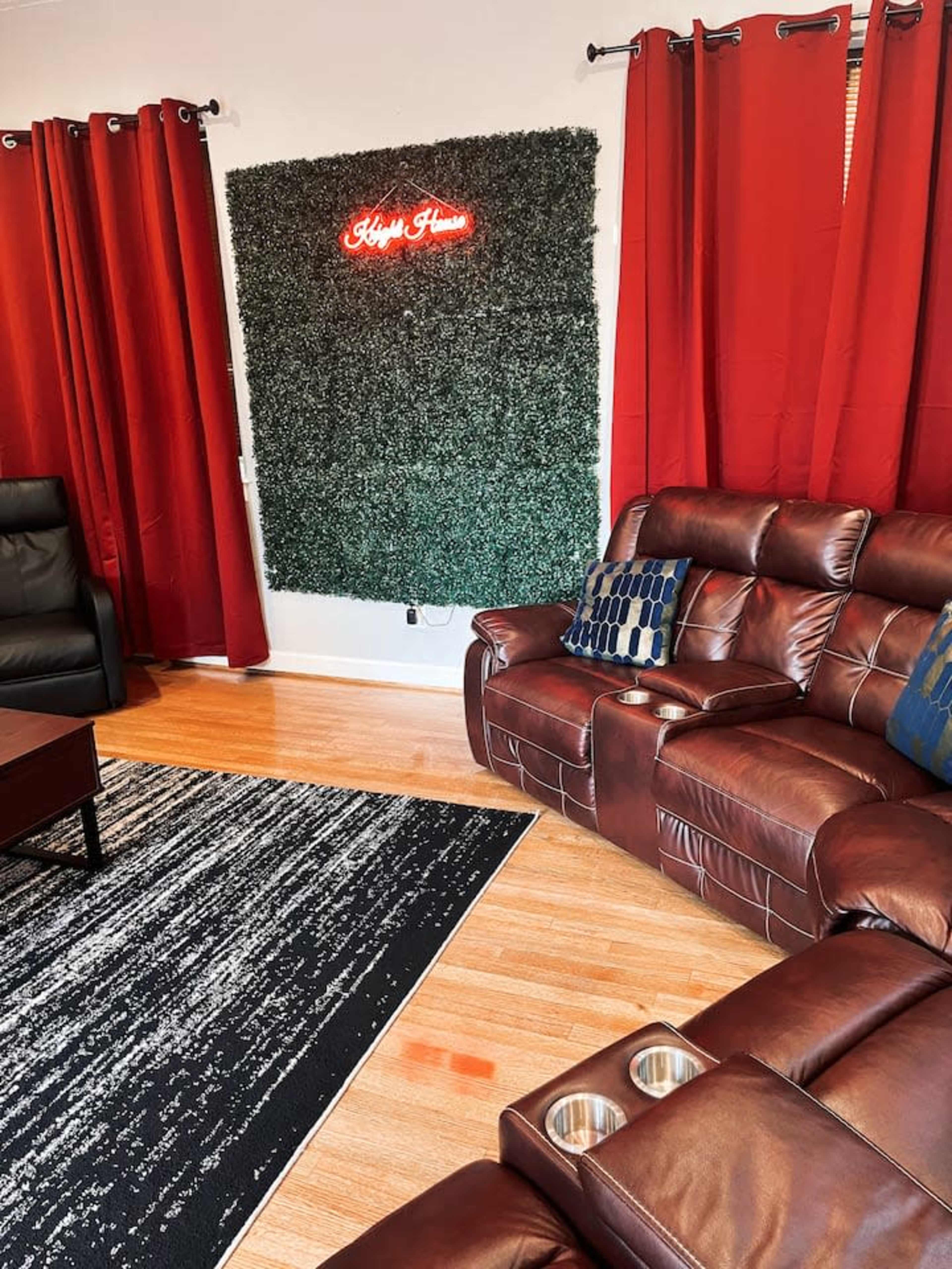 A living room with dark red curtains, a green plant wall, a brown sectional sofa, and a coffee table on a patterned rug.