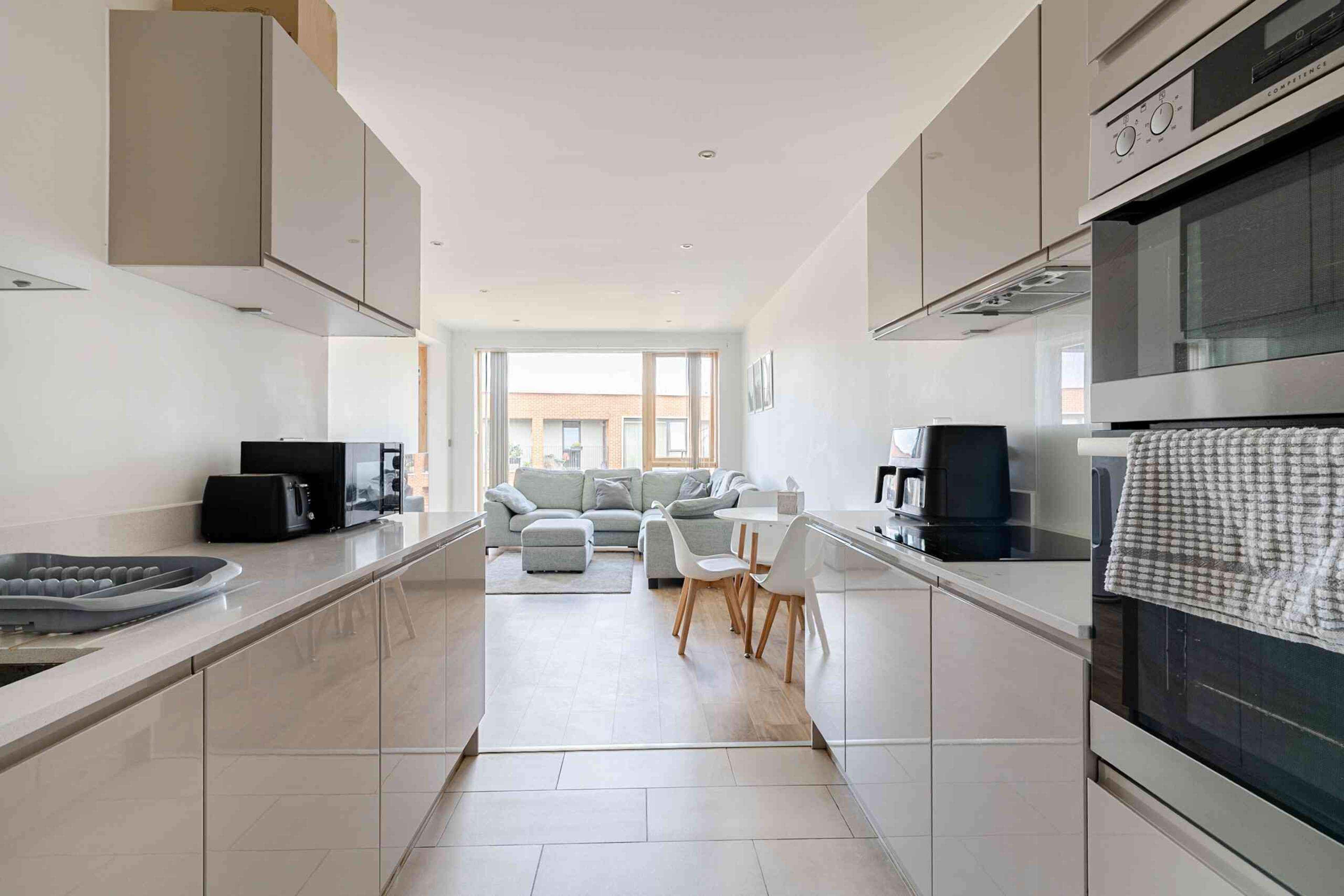 A modern kitchen with sleek cabinets and appliances opens into a light-filled living area furnished with a sofa and dining table.