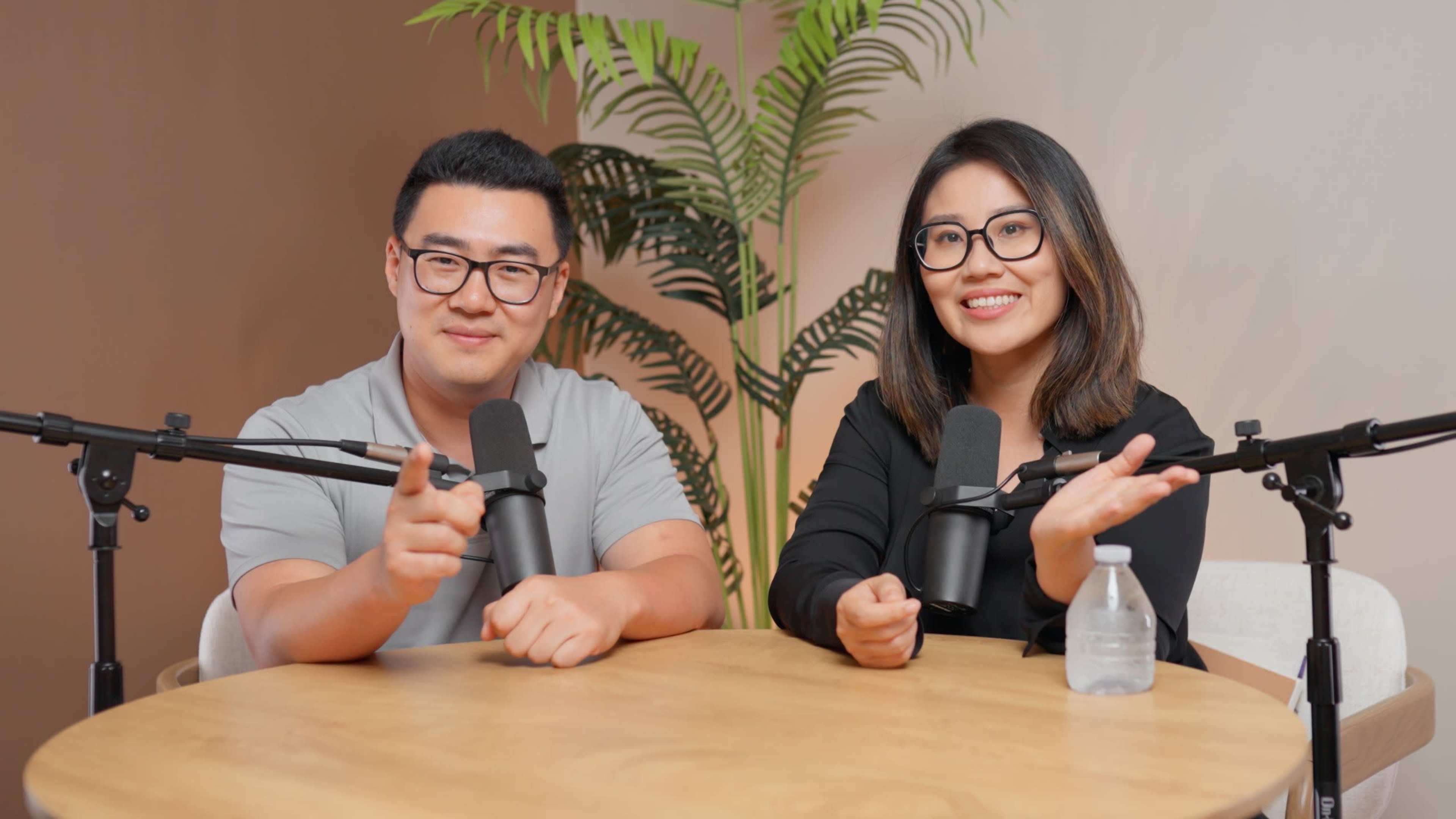 Two individuals with glasses are seated at a round wooden table with microphones in front of them, smiling and gesturing, while a small water bottle is placed on the table.