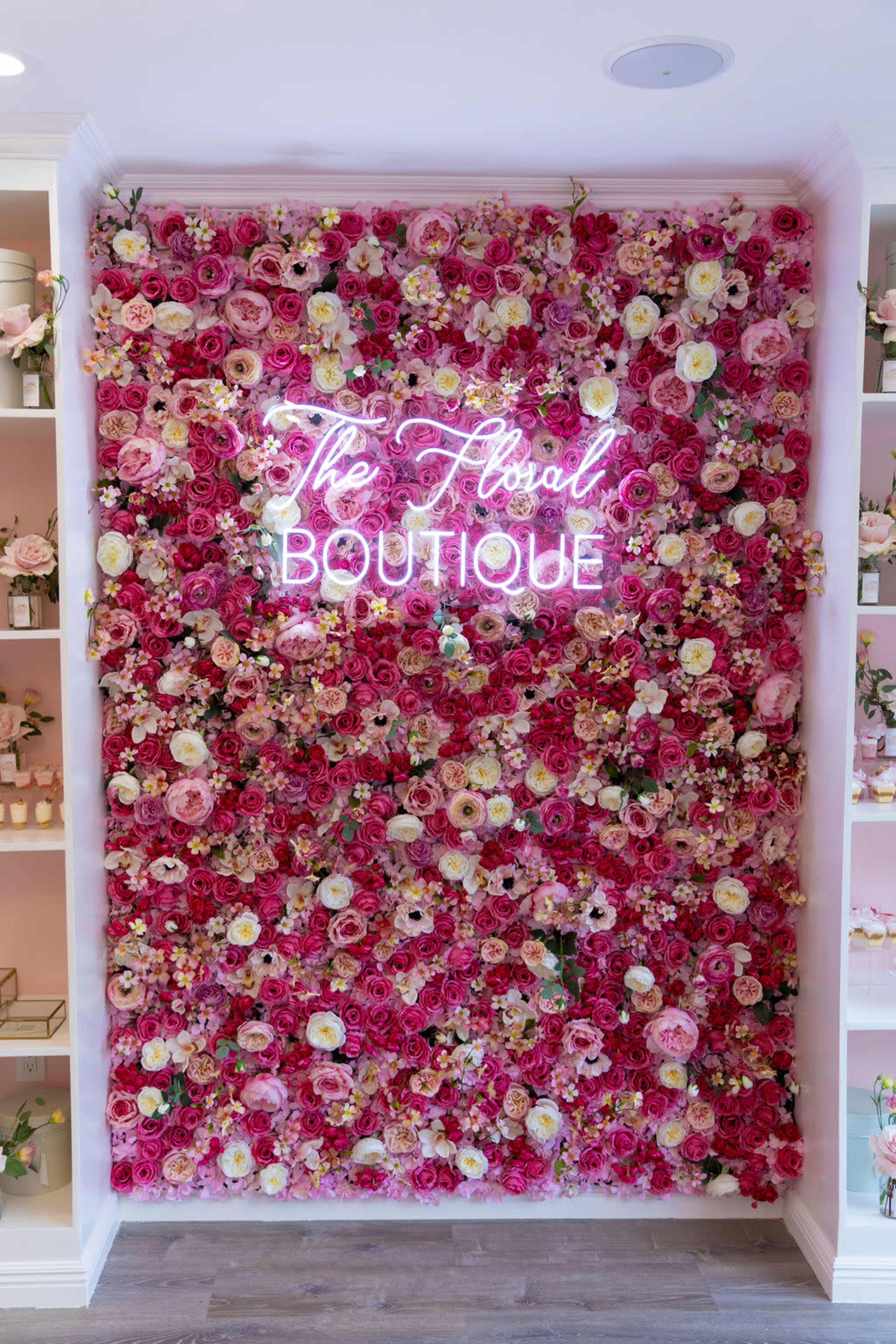 The image shows a wall decorated with an arrangement of pink and cream flowers, featuring a neon sign that reads "The Floral Boutique."