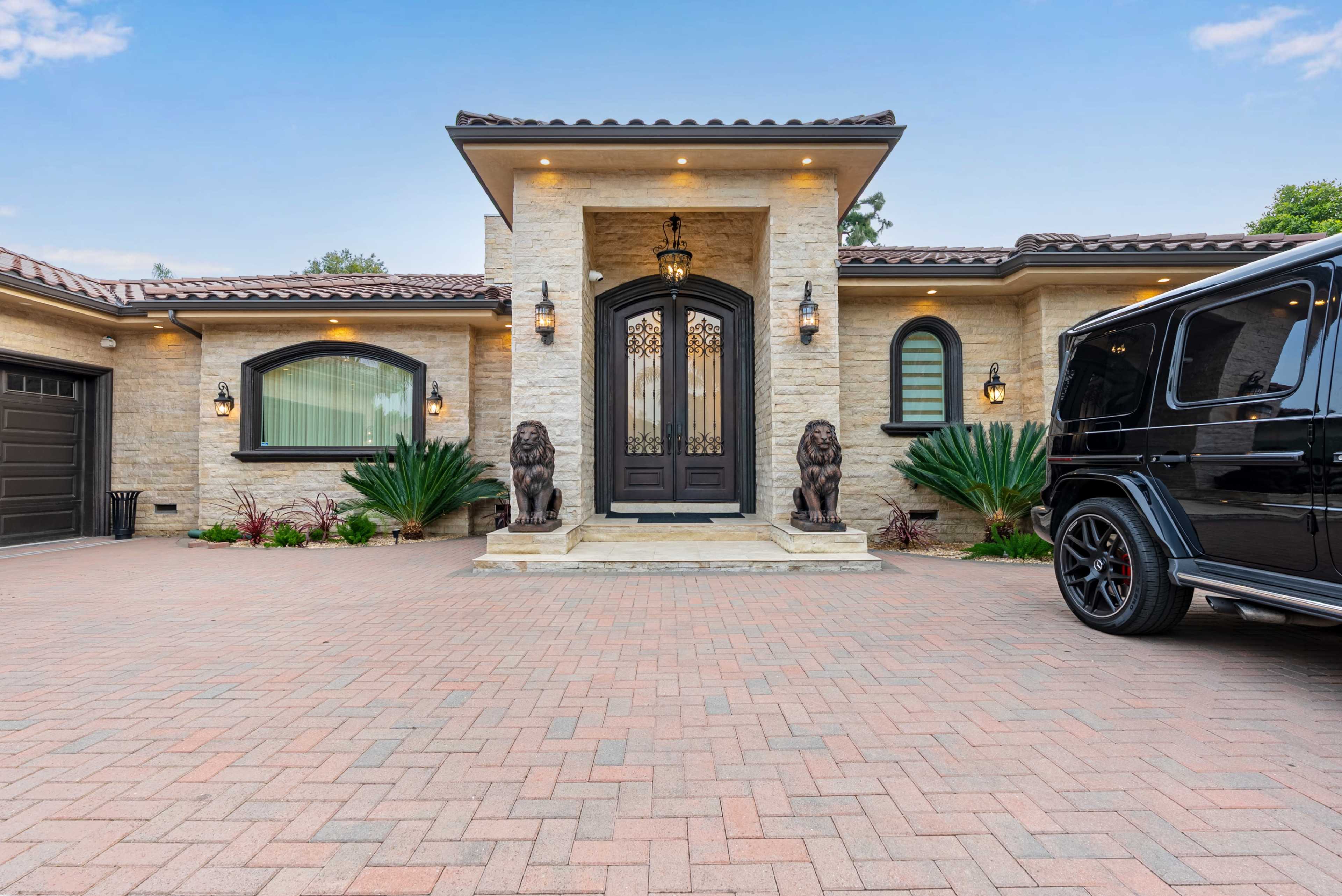 The image shows a luxurious house featuring a stone facade, a grand entrance flanked by two lion statues, and a paved driveway with a black SUV parked on the side.
