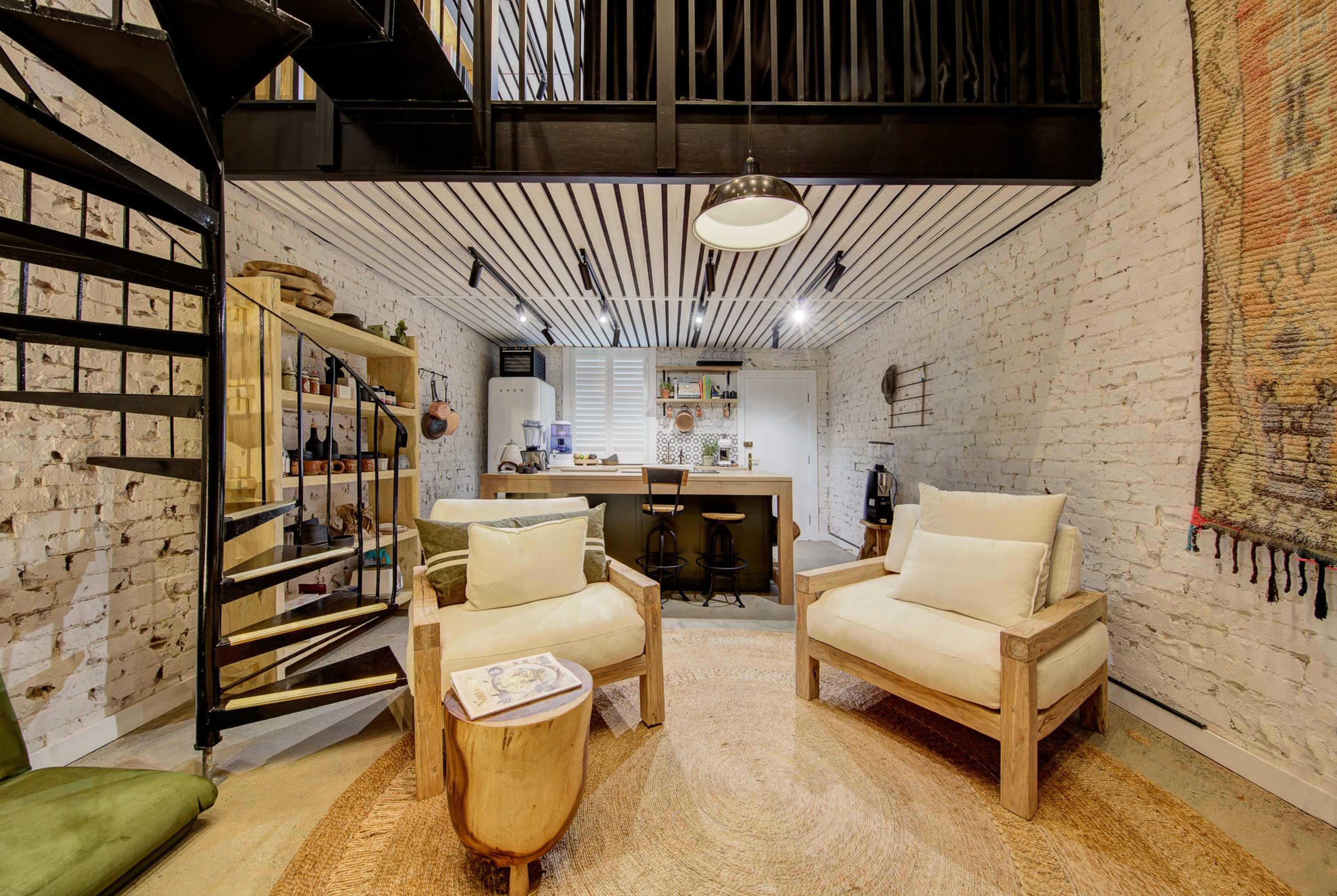 The image depicts a modern interior space with a spiral staircase, two wooden chairs, and a kitchen area, featuring exposed brick walls and a round rug.