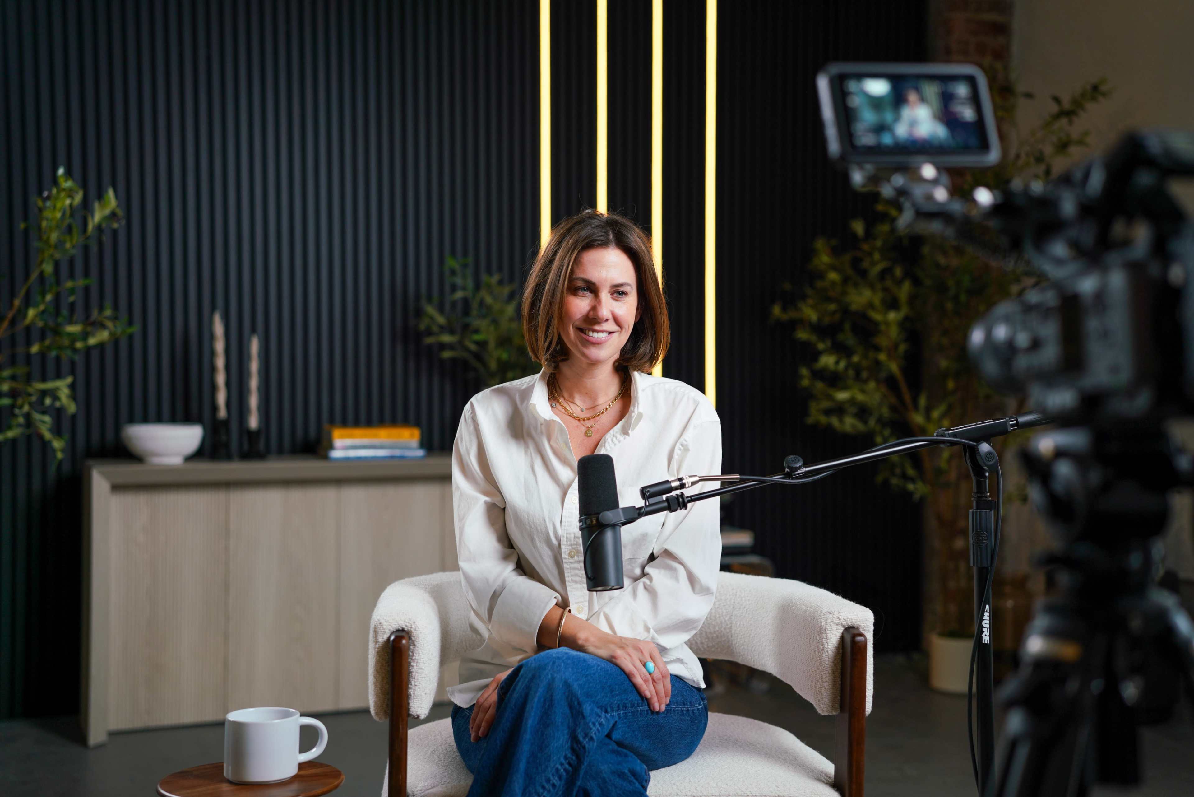A woman sits in a modern studio setup with a microphone and camera, smiling while positioned in front of a wooden table and a backdrop of vertical stripes.