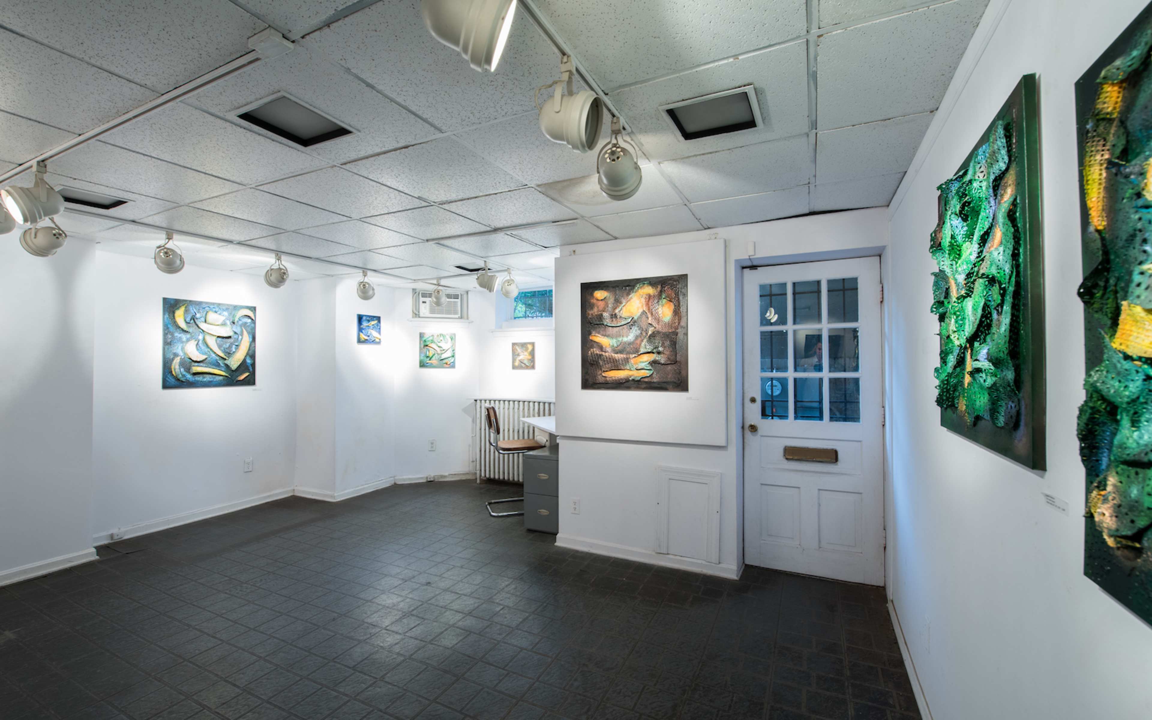 An art gallery with artworks displayed on white walls, a door on the right, and a desk in the corner.