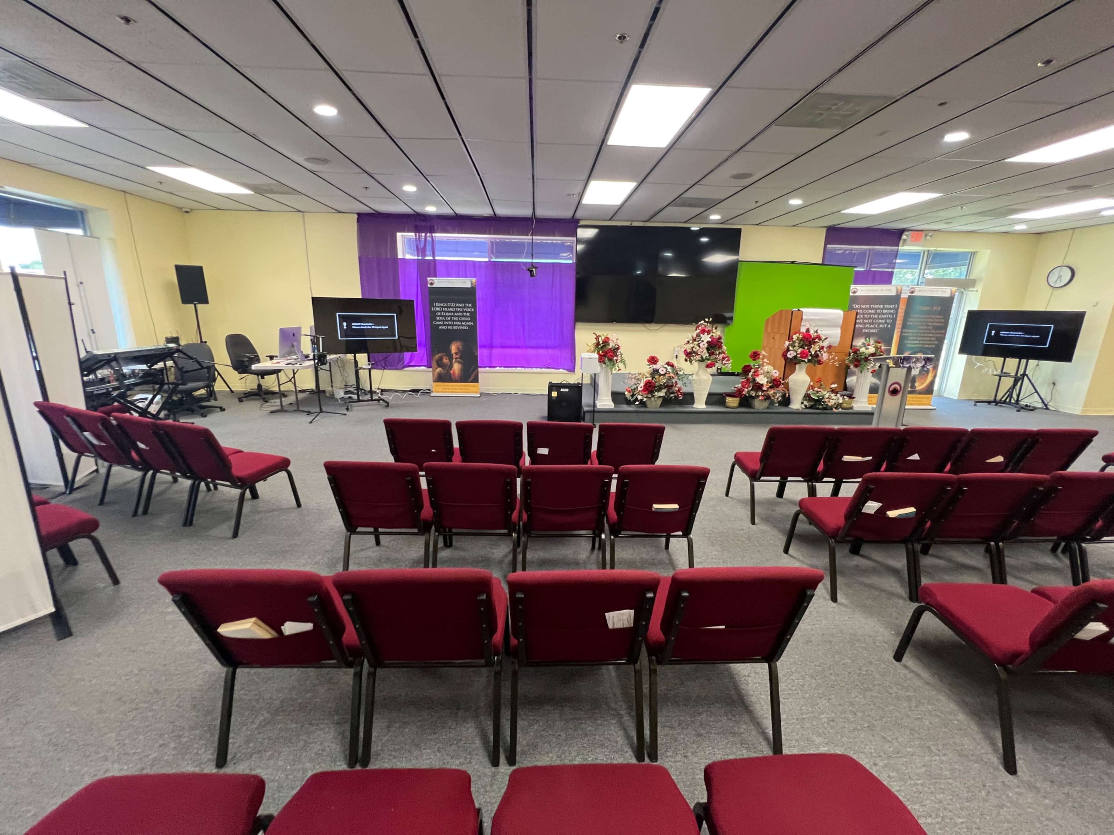The image shows a room set up for an event, featuring rows of red chairs facing a stage with floral arrangements, screens, and a green backdrop.