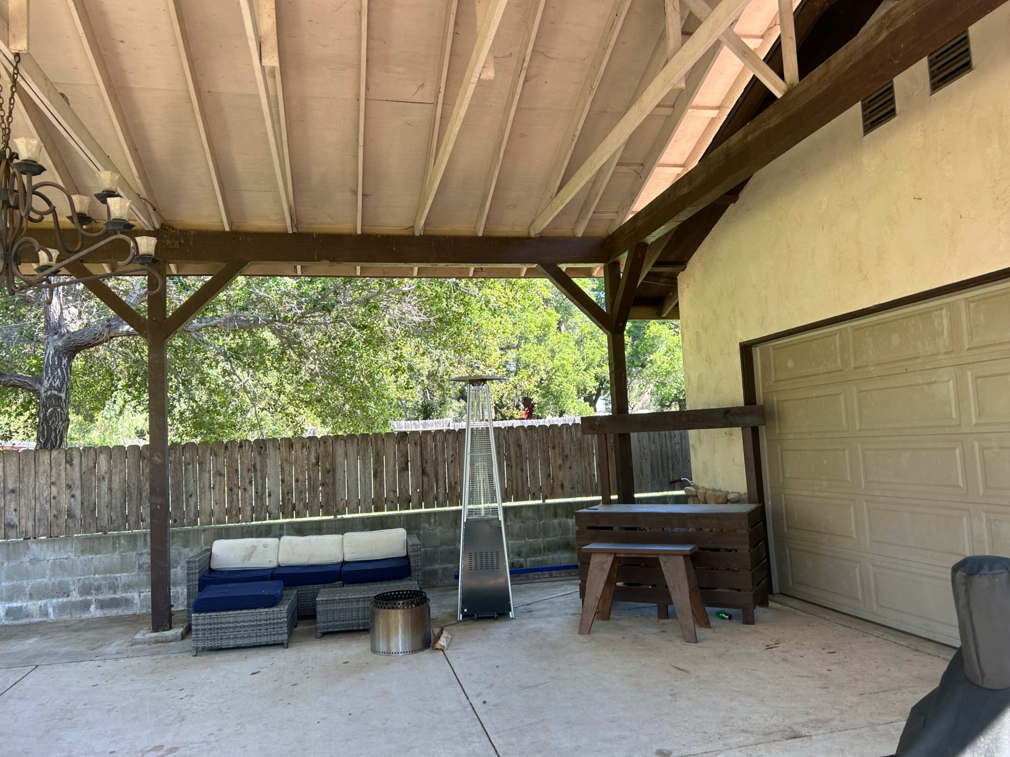 The image shows a covered patio area with a sofa set, a heater, and a wooden table, adjacent to a garage door and surrounded by a wooden fence.