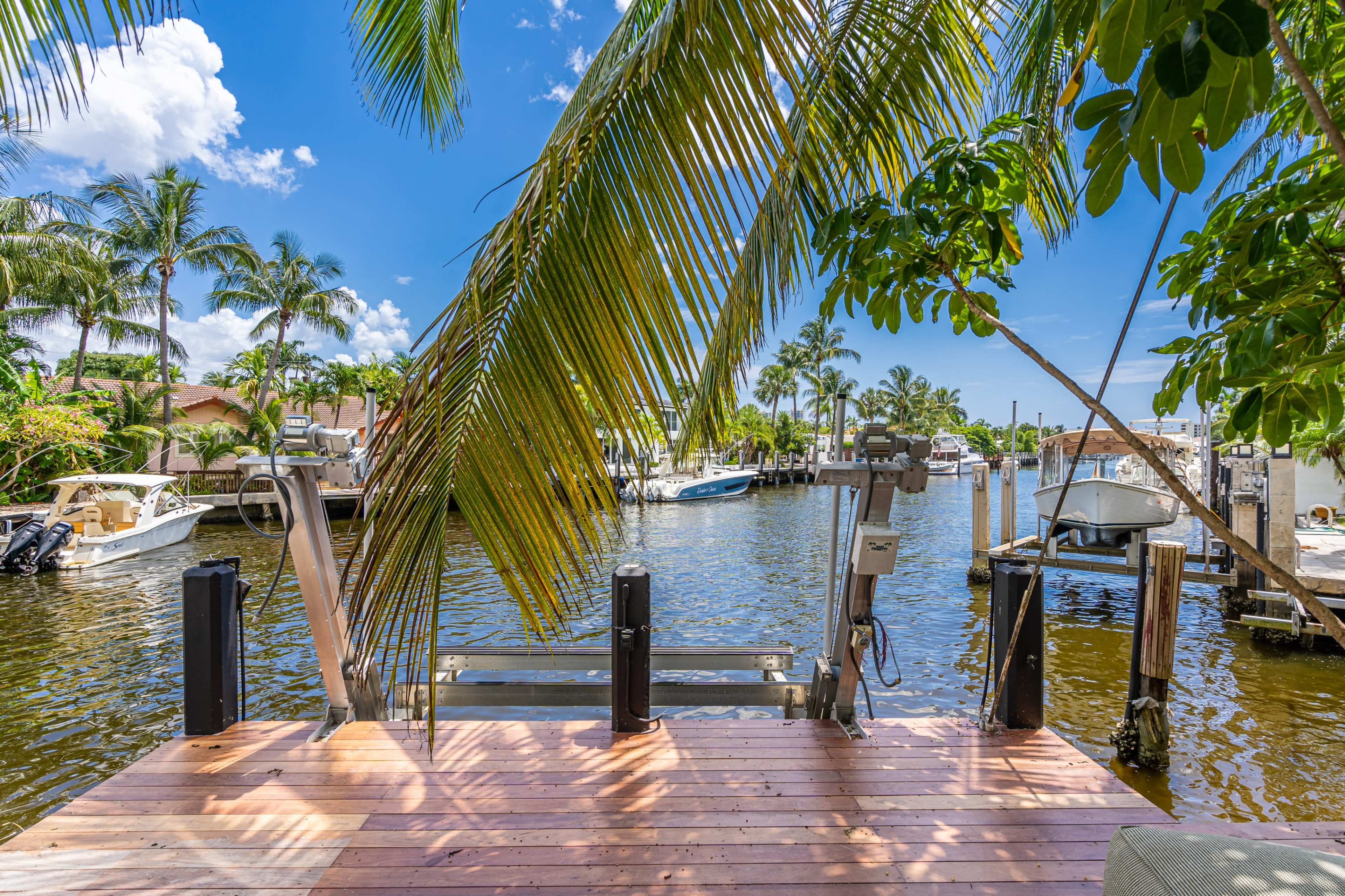 A wooden dock extends over a canal lined with boats and palm trees under a clear blue sky.