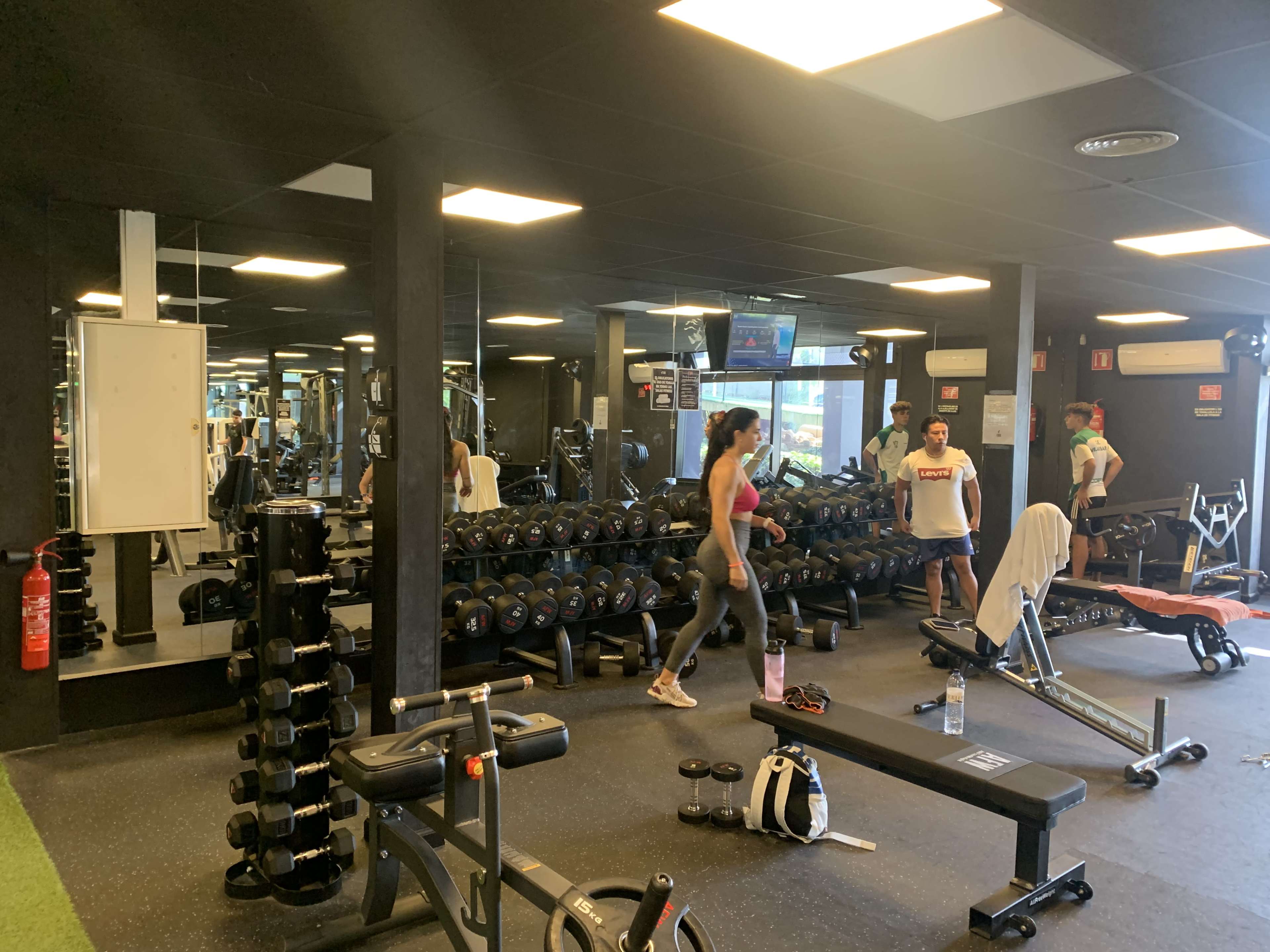 A gym interior with several weightlifting benches, dumbbells, and people exercising in various areas.
