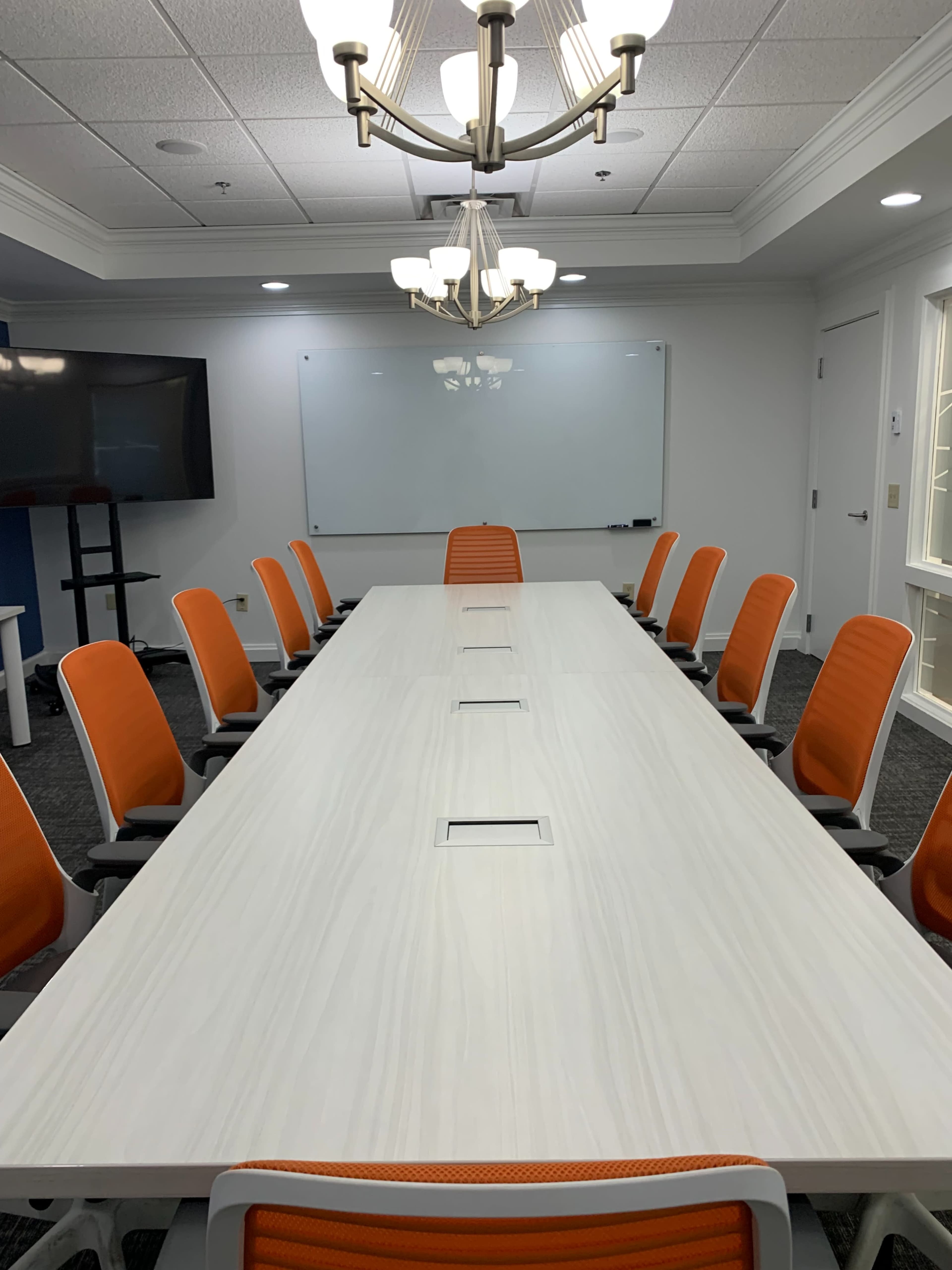 A long conference table with orange chairs is set up in a well-lit meeting room, featuring a whiteboard and a large screen.