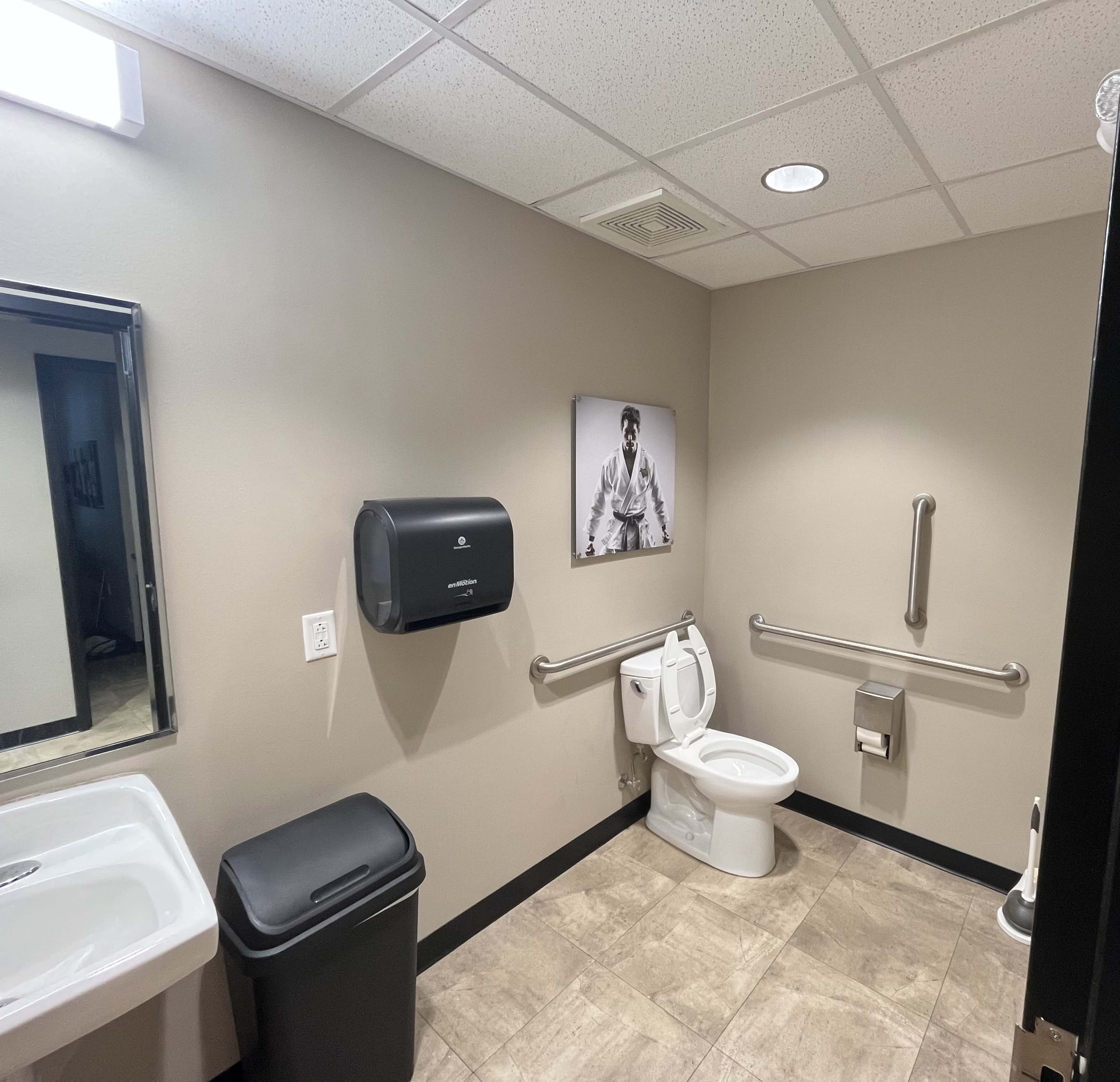 The image shows a restroom with a toilet, sink, trash can, and a wall-mounted paper towel dispenser, featuring a black-and-white photograph on the wall.