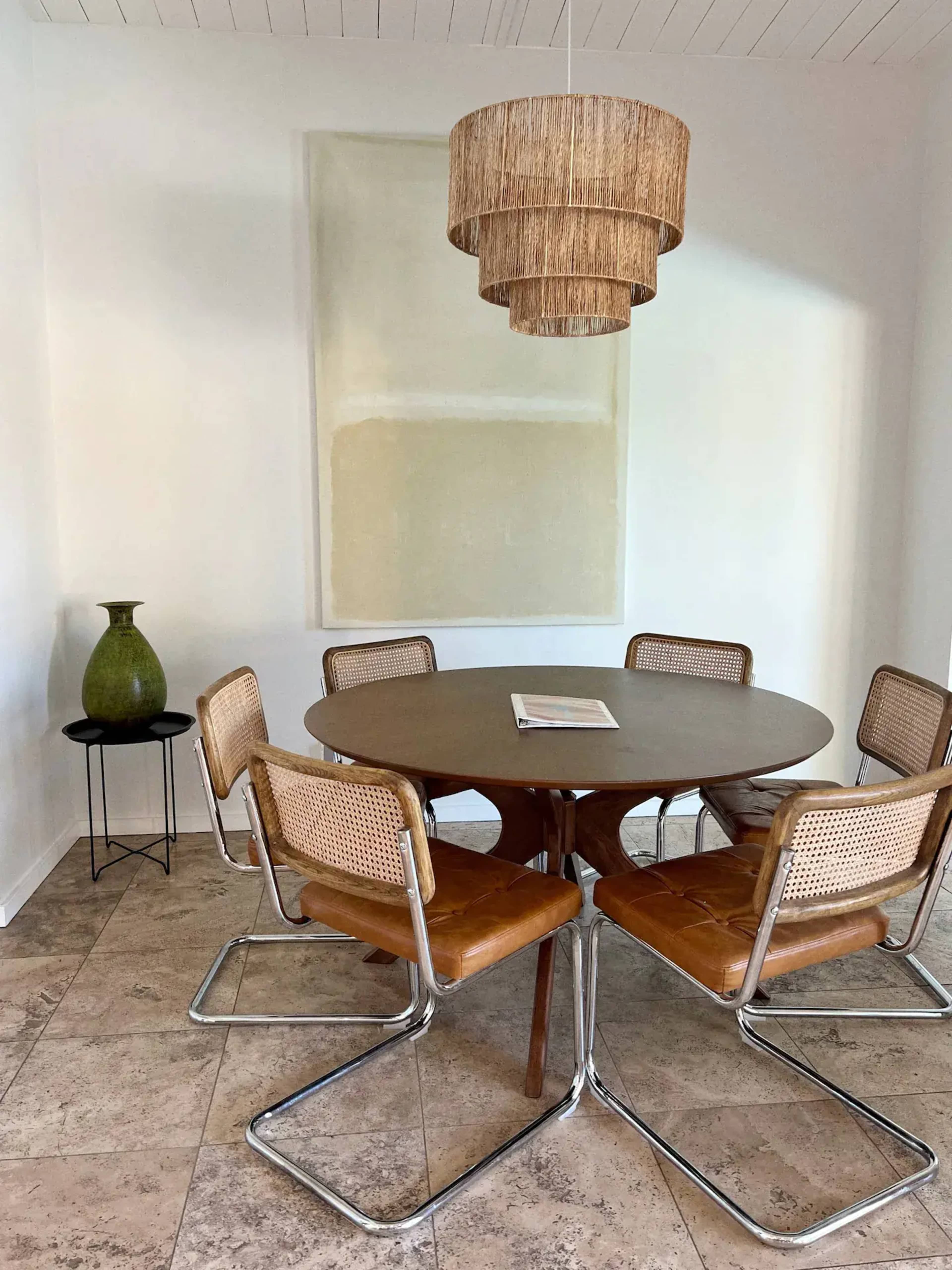 A round wooden dining table surrounded by six leather and metal chairs is placed in a light-filled room with a large woven pendant light and a green vase on a small side table.