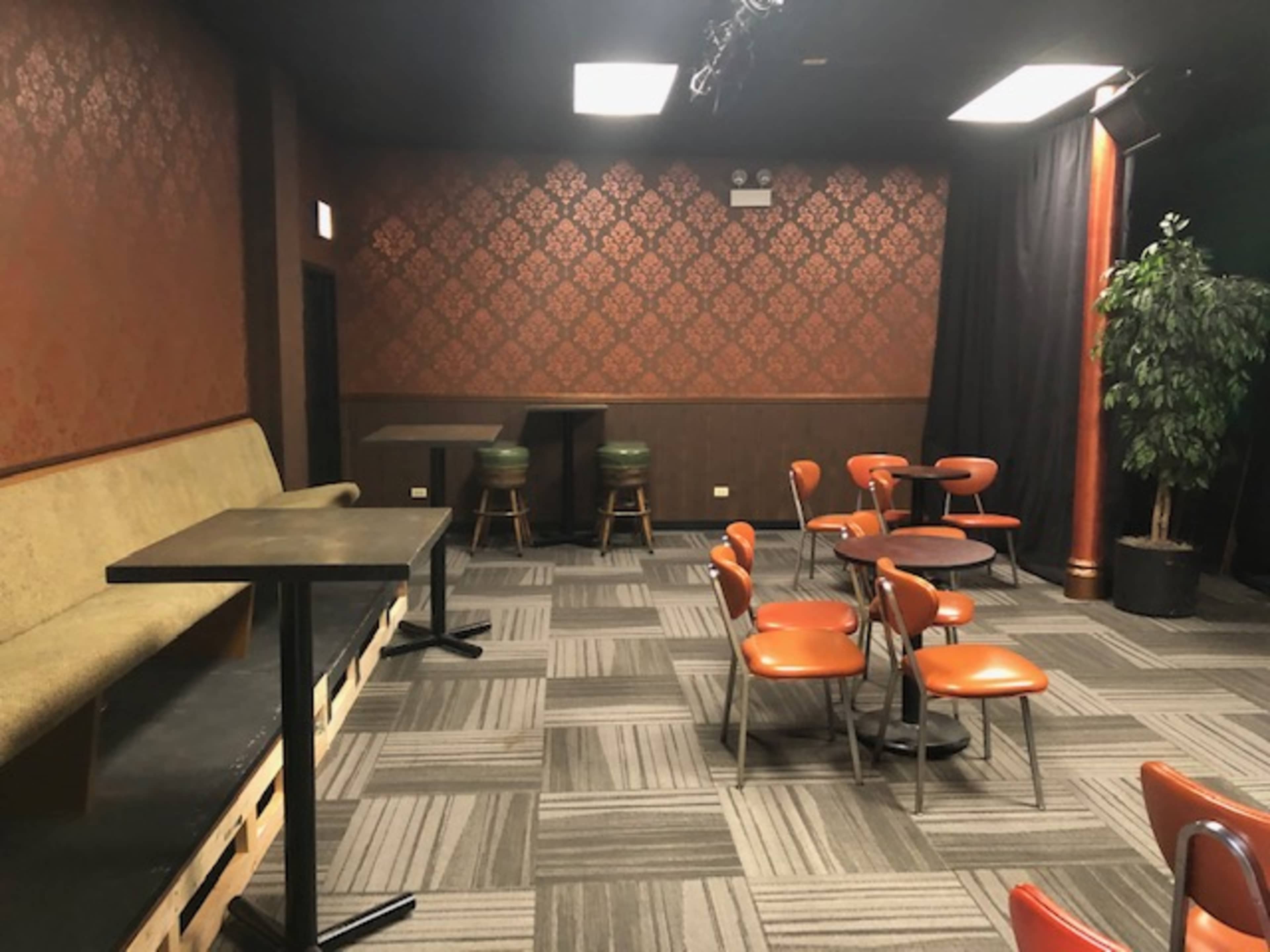 An empty performance space with patterned wallpaper, a seating area with a long couch, round tables, and orange chairs.