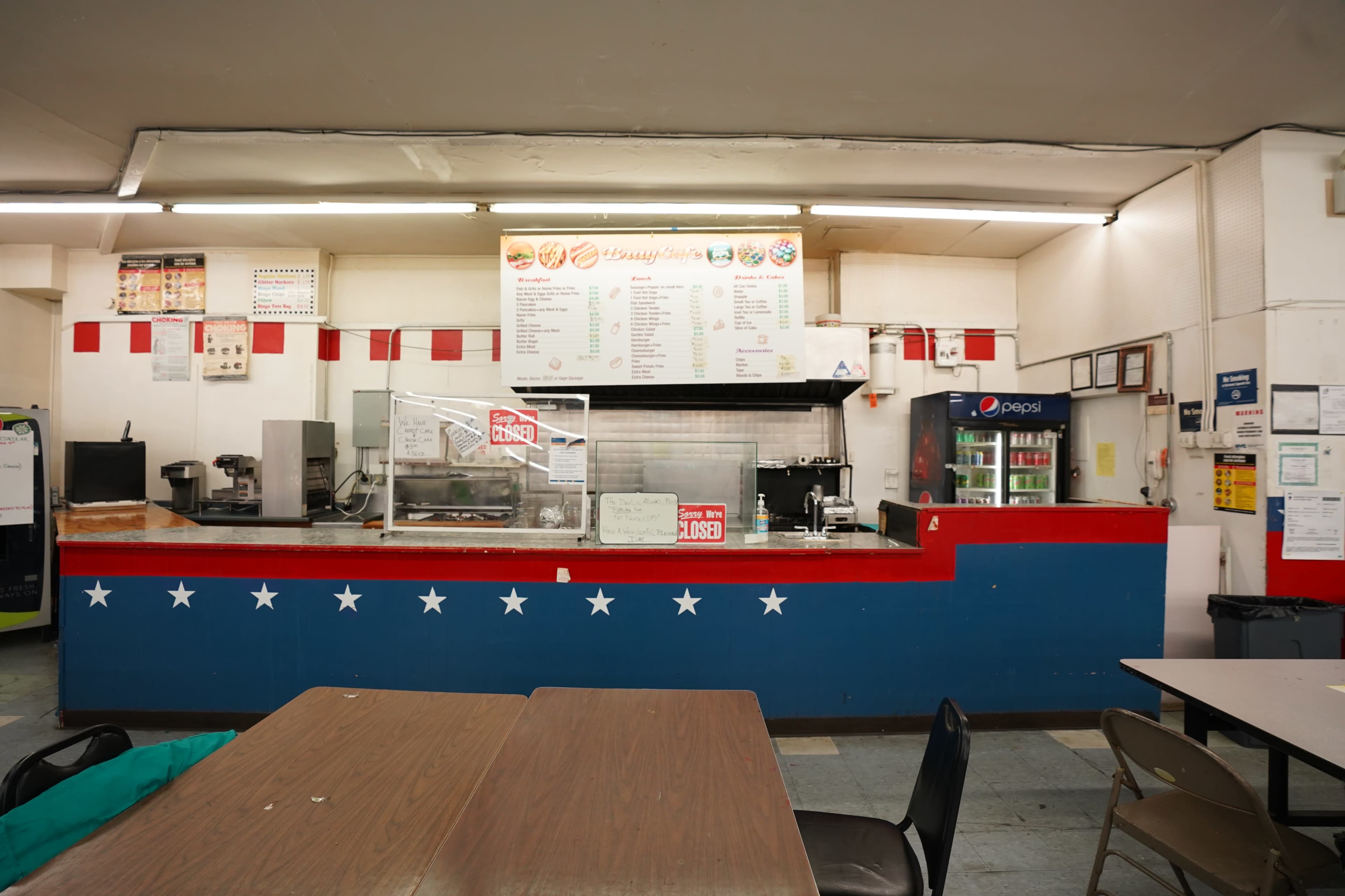 The image shows a food counter in a casual dining area, featuring a menu display, a drink cooler, and tables with chairs nearby.