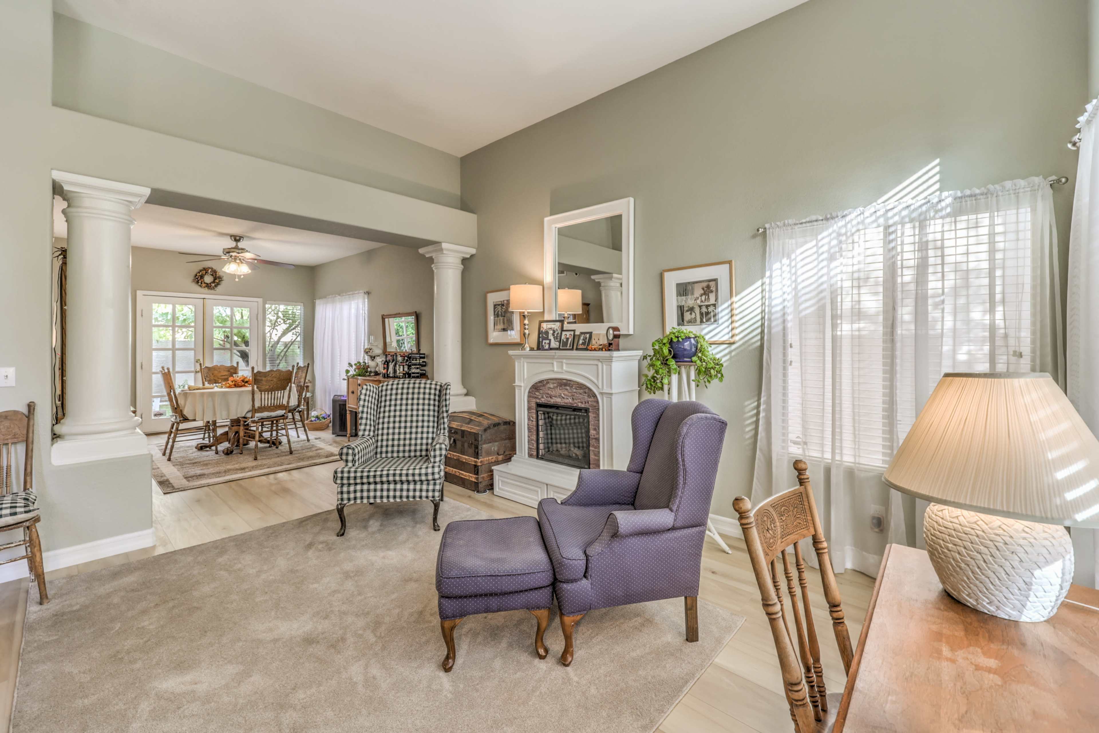 A living room features two patterned armchairs facing a fireplace, with a view into a dining area through an archway.