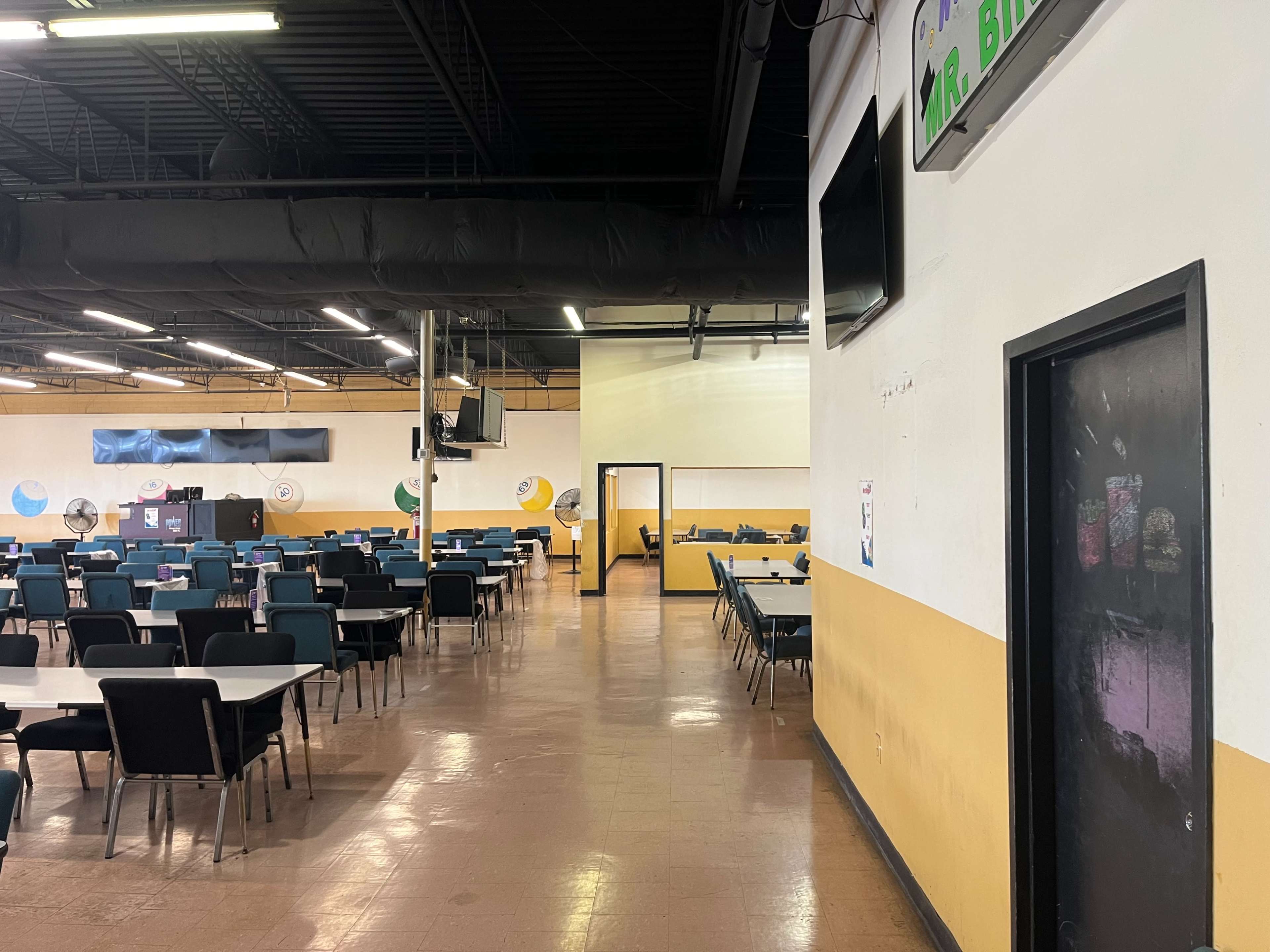 A spacious dining area with rows of tables and chairs, leading to a doorway on the right and a kitchen or service area in the background.