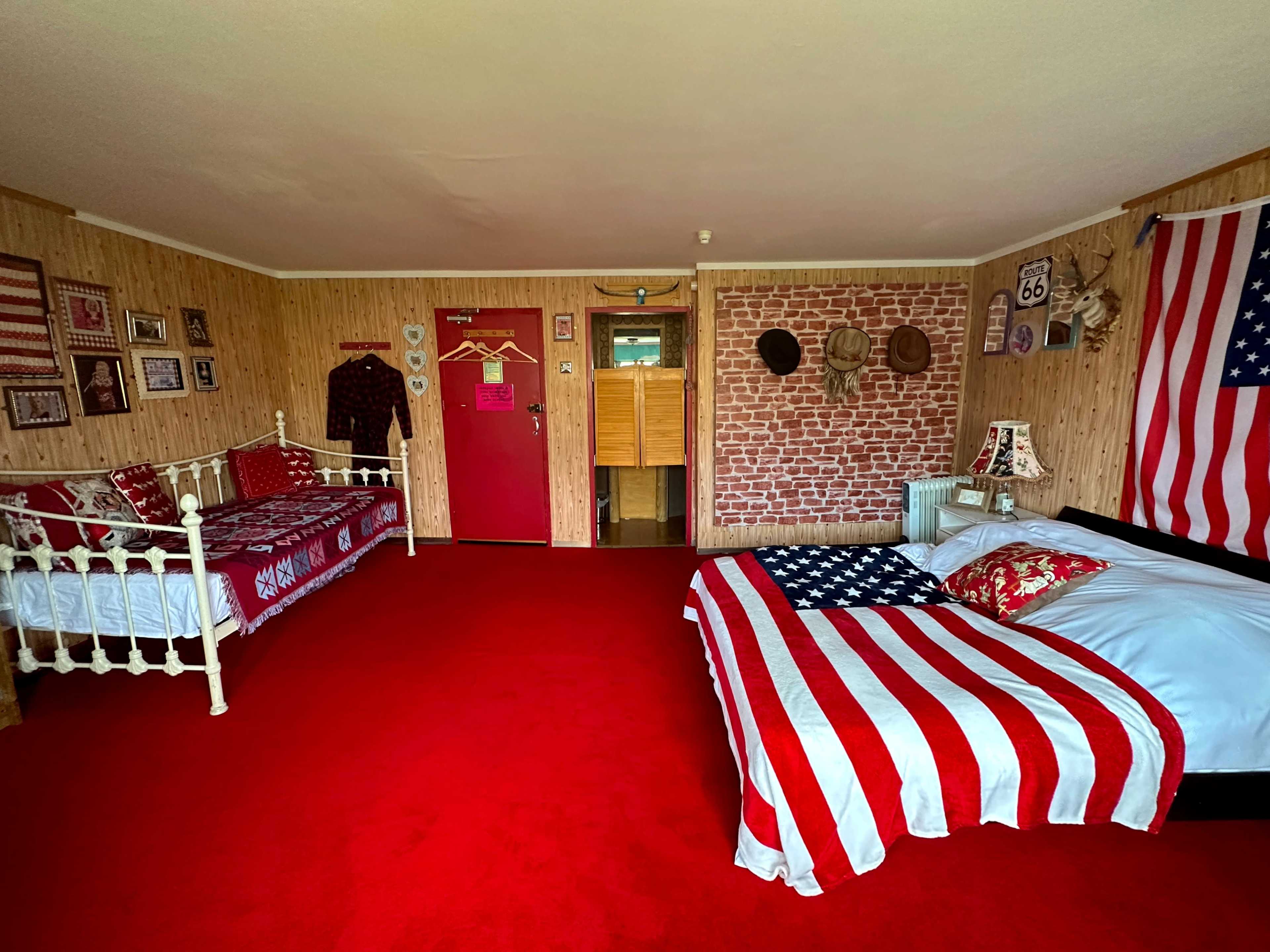 The room features two beds, one with an American flag bedspread, red carpet, and walls adorned with patriotic decor and vintage items.
