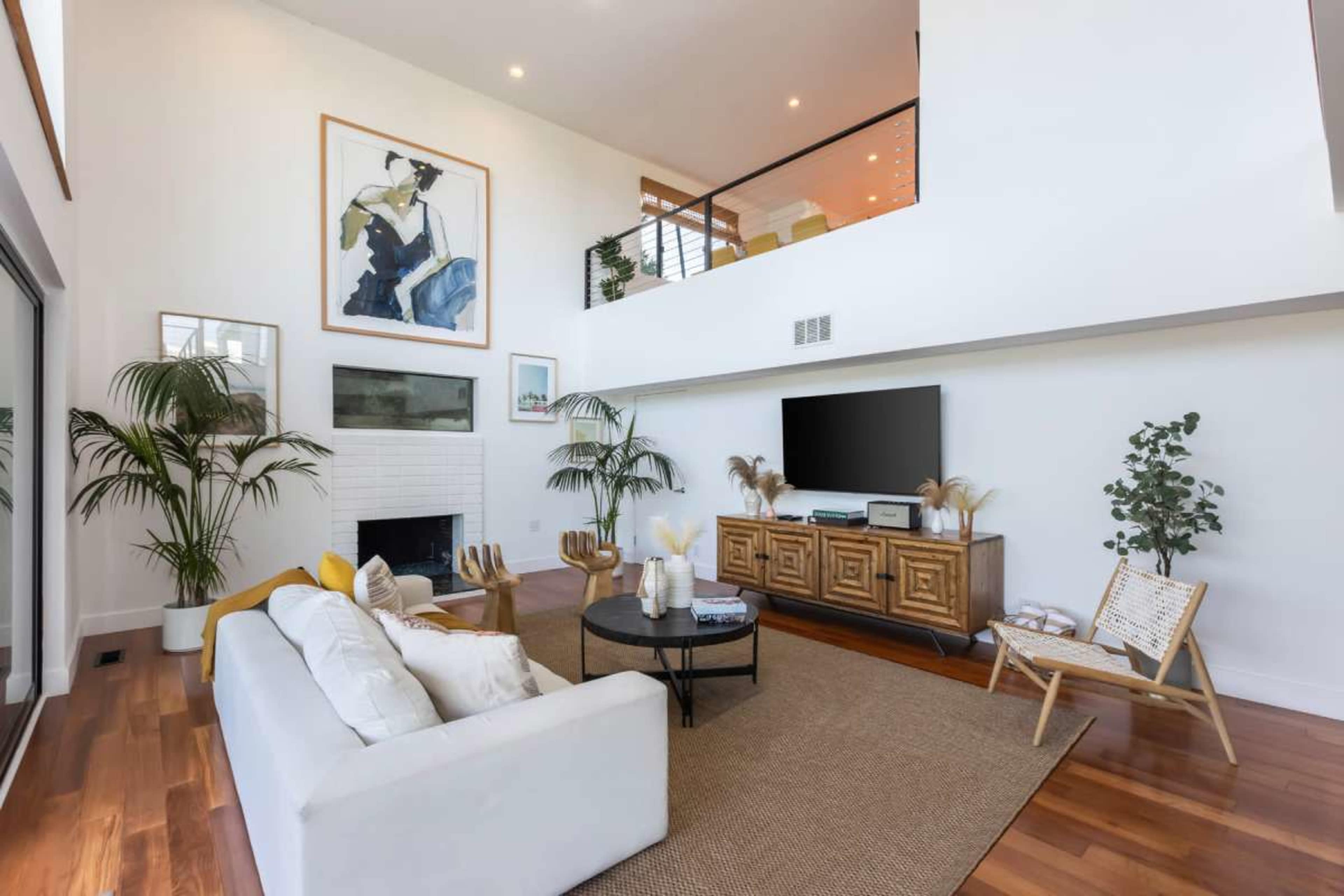 A modern living room features a white sofa, a round coffee table, and a large TV mounted on a wall, with plants and artwork complementing the space.