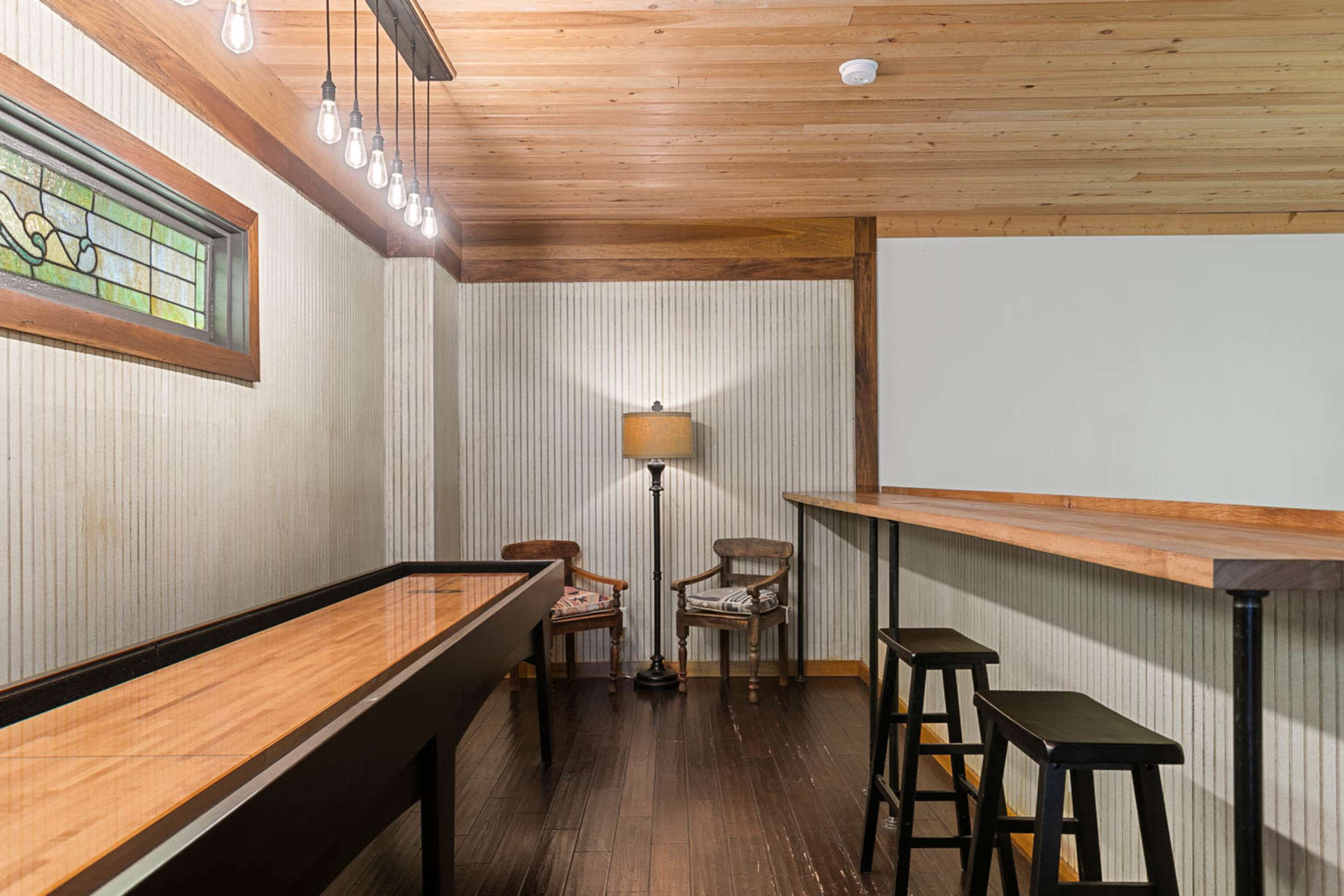 The image shows a neatly designed room featuring a wooden bar top, two black stools, a lamp, and two chairs, all situated under a ceiling with exposed bulbs and a stained glass window.