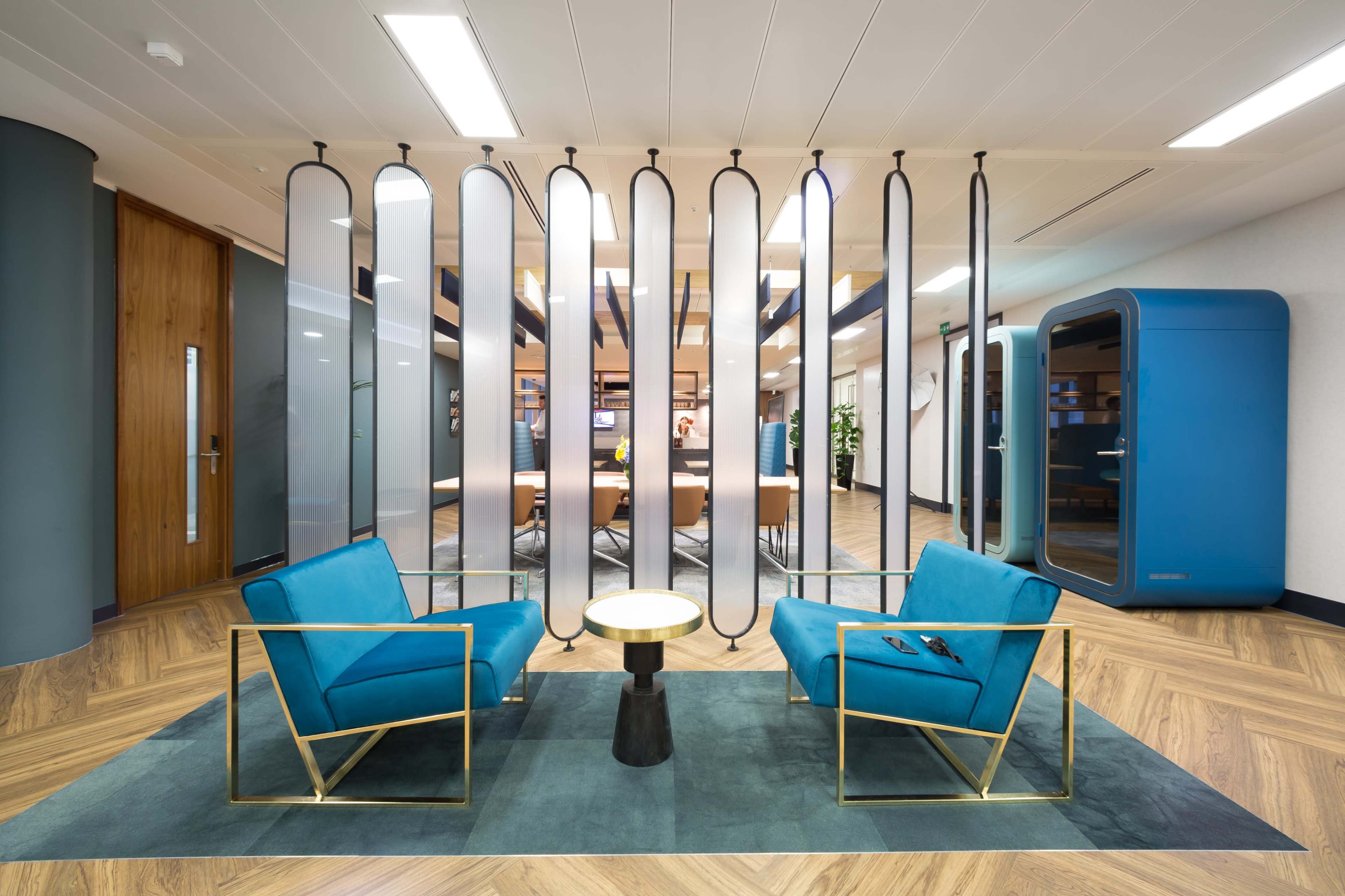 The image shows a modern office lounge area with two teal chairs and a small round table, separated by a decorative screen.