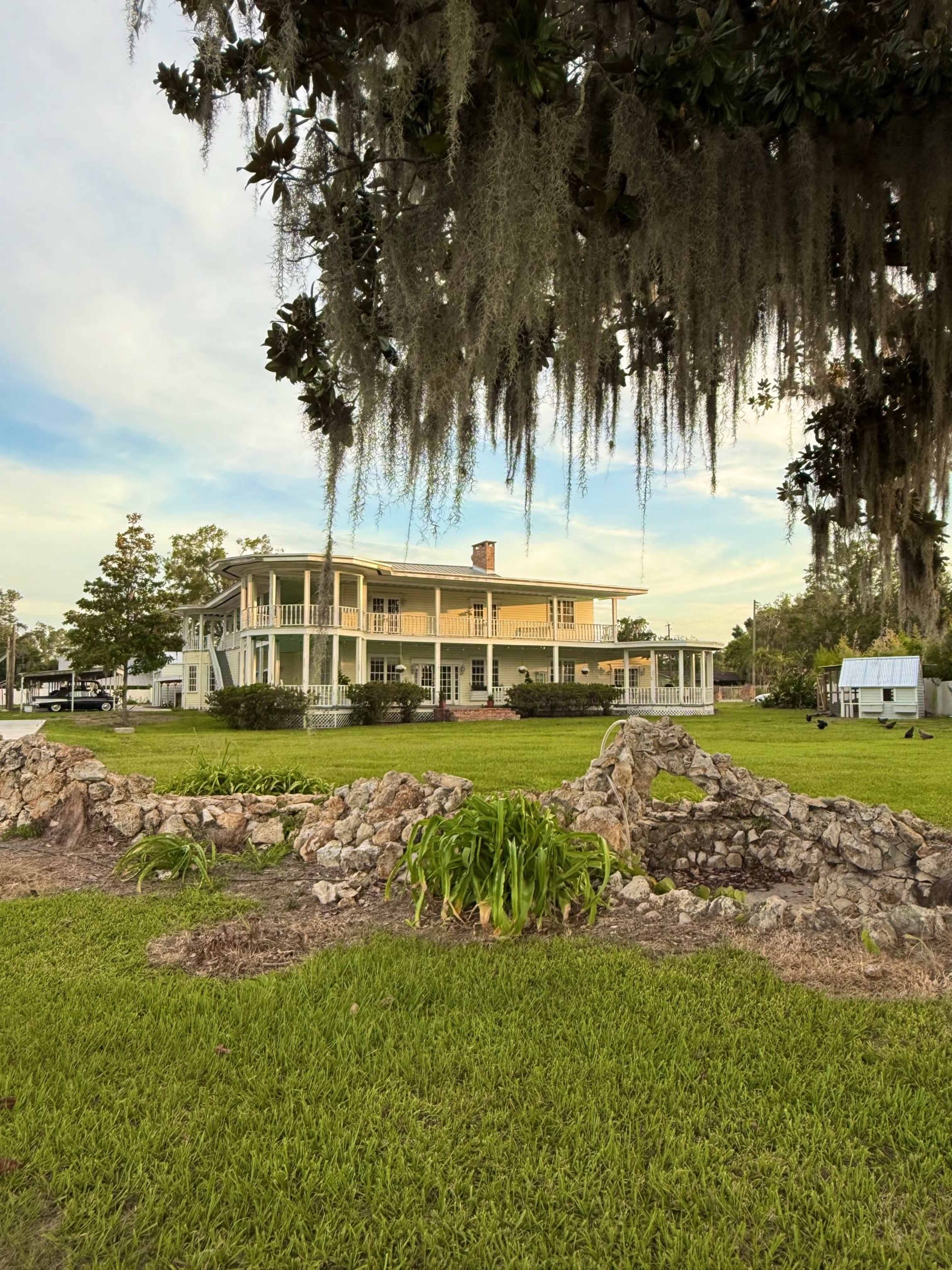 A large, two-story house with a wraparound porch sits behind a stone garden wall, surrounded by greenery and hanging moss.