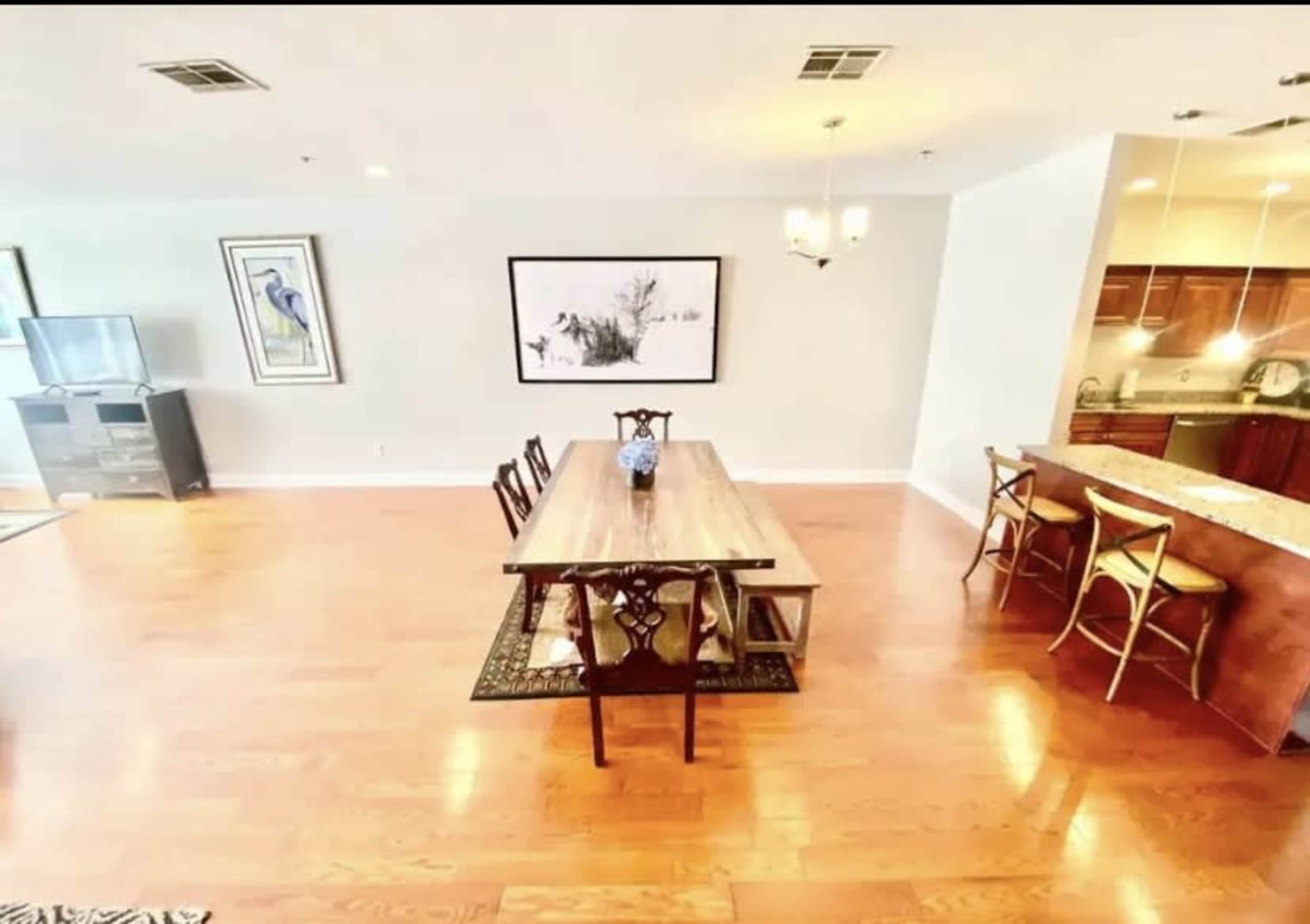 The image shows a spacious dining area with a long wooden table, surrounded by chairs, and an open kitchen visible in the background.