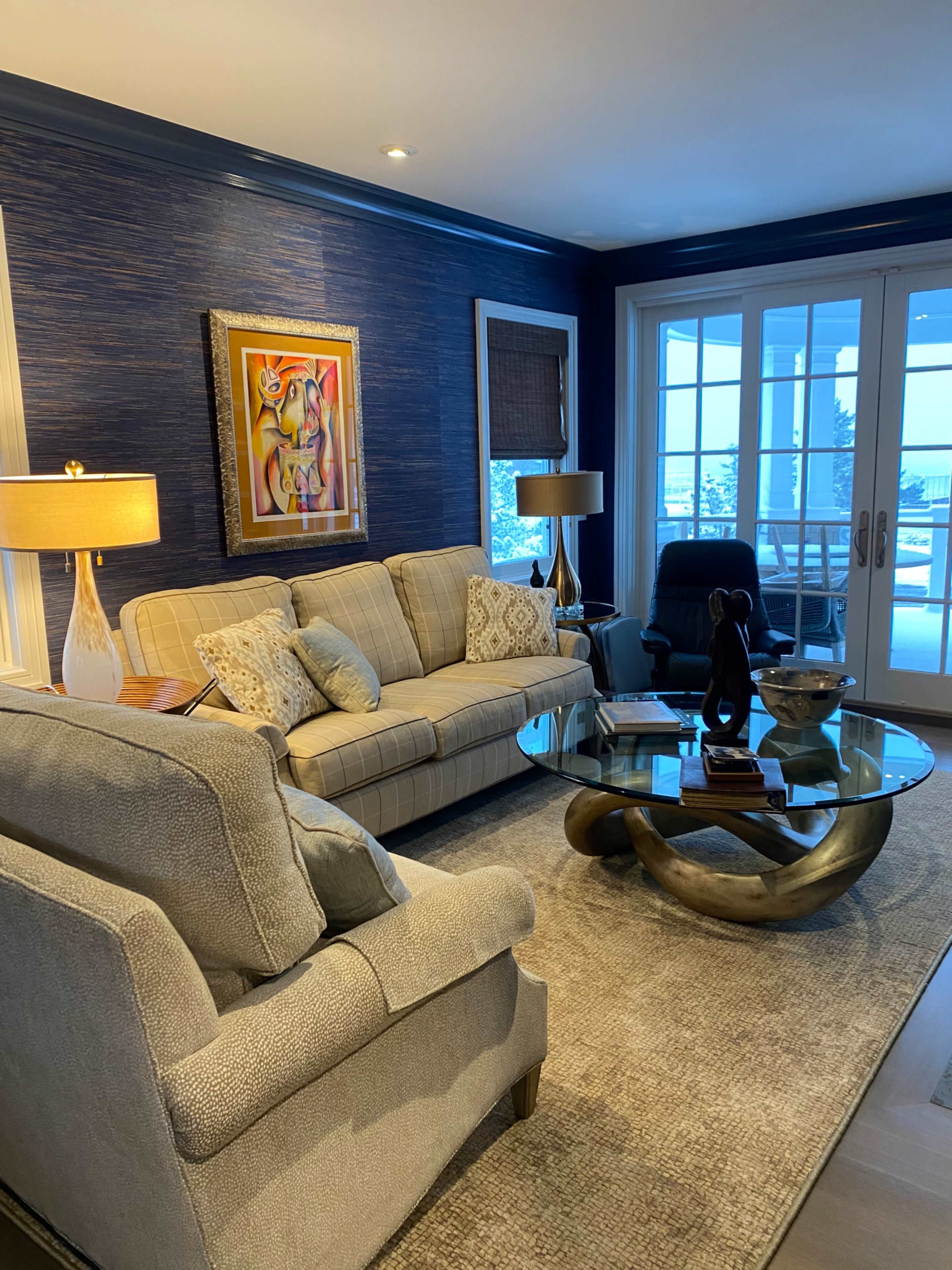 The image shows a stylish living room featuring a light-colored sofa, a matching armchair, a glass coffee table, and large windows with sliding doors.