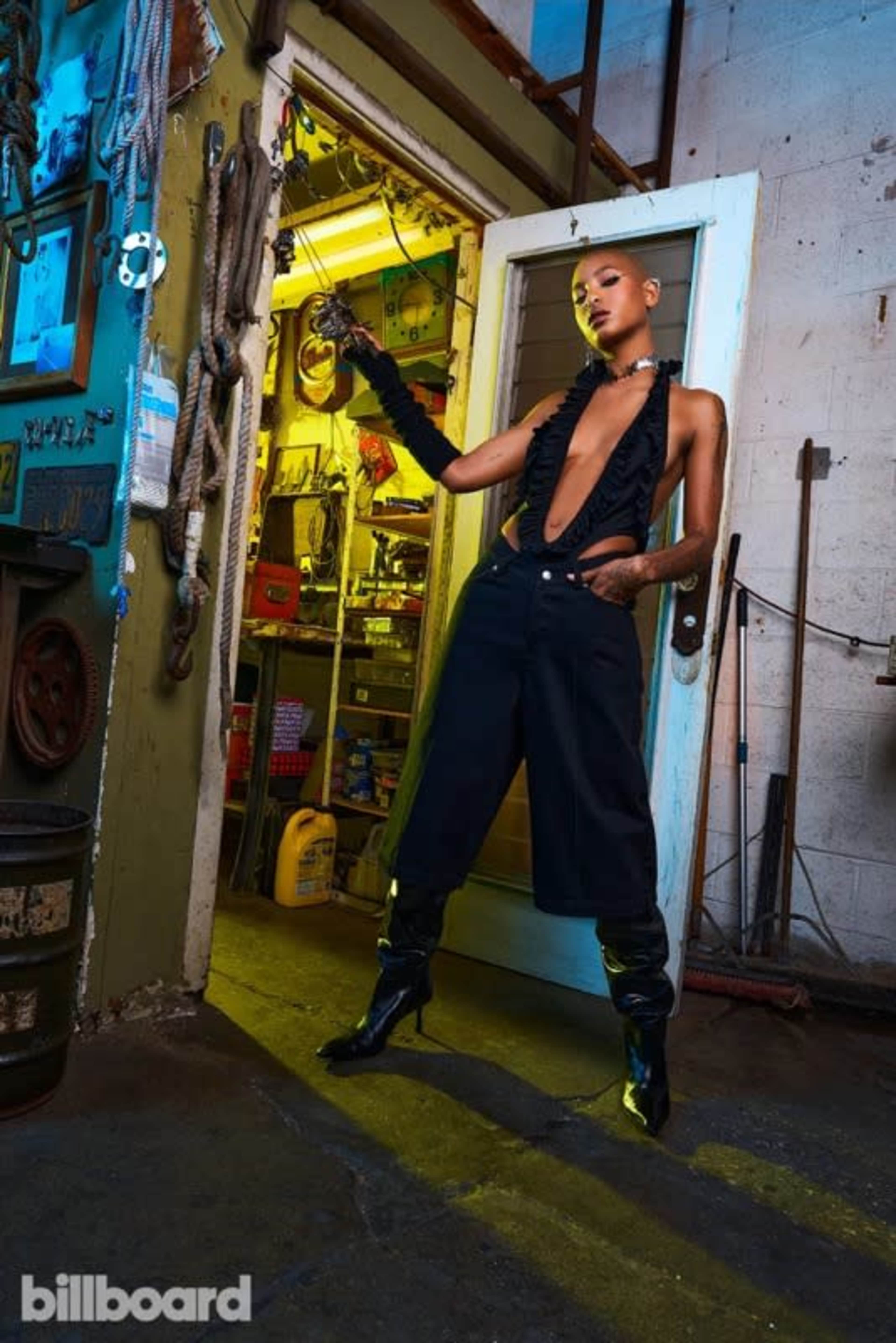 A model poses in a dimly lit industrial space, wearing a bold outfit with a deep neckline and high-waisted pants.