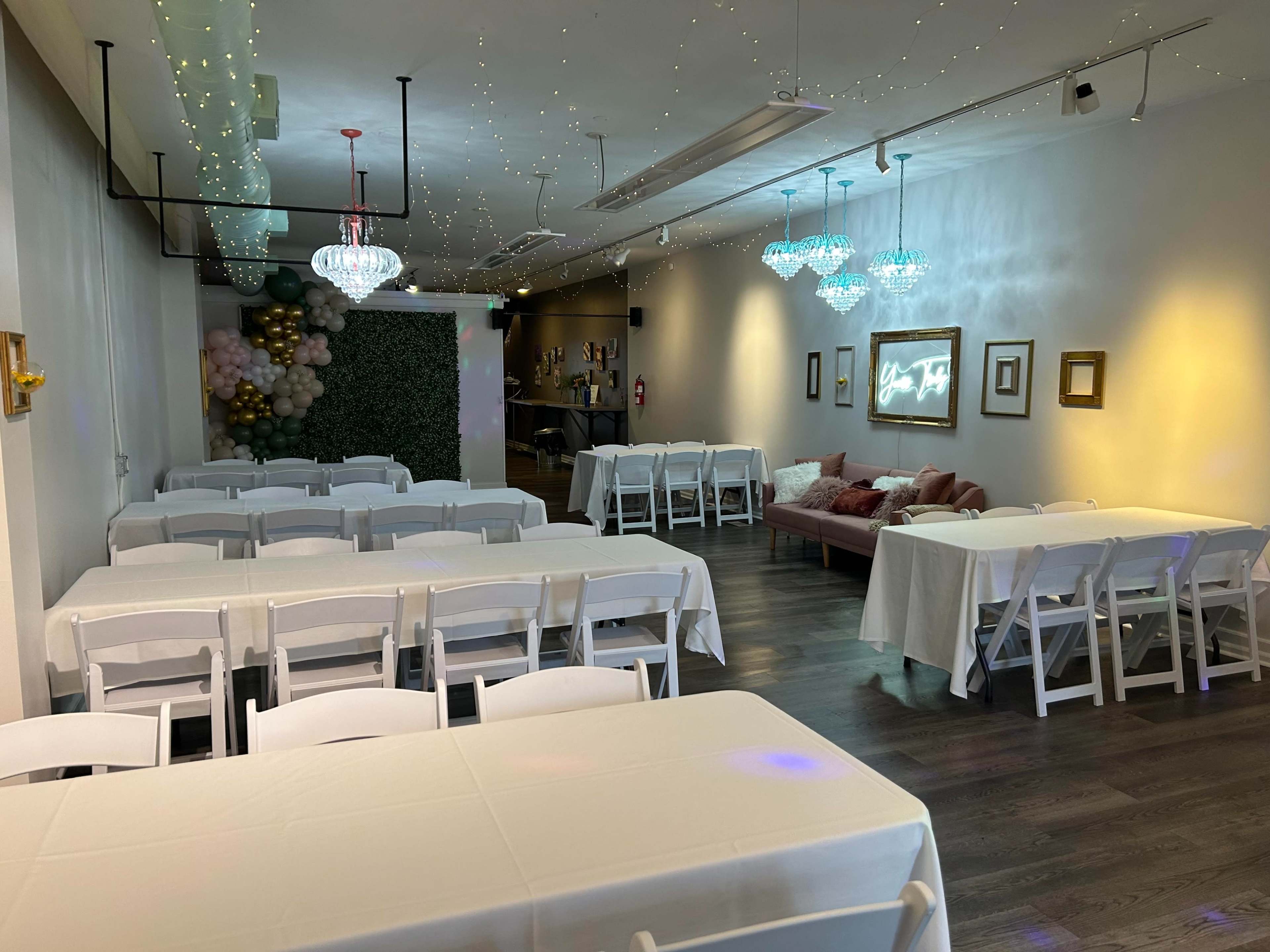The image shows an indoor event space with rows of tables covered in white cloth, decorated with string lights, and featuring a lounge area with a pink couch.